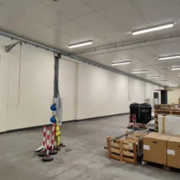 Wnętrze hali w trakcie prac instalacyjnych: widoczne kable w korytkach, lampy sufitowe, elementy zabezpieczeń i materiały budowlane na paletach, betonowa posadzka.