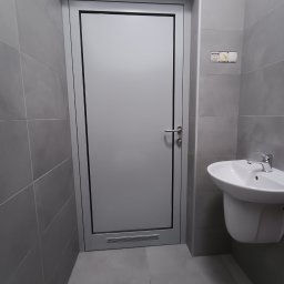 PERFECT-BUD - Szara łazienka z umywalką i drzwiami. Płytki na ścianach i podłodze w odcieniach szarości. Minimalistyczny design, widoczne gniazdka elektryczne.