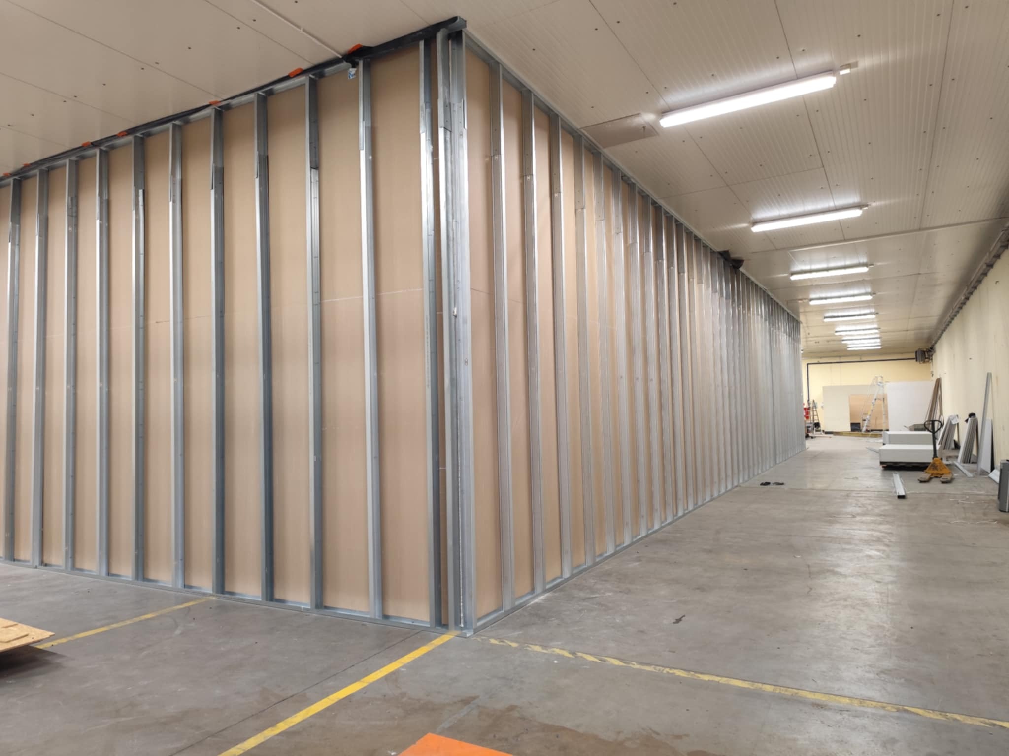 Konstrukcja ścian działowych z płyt kartonowo-gipsowych na metalowym stelażu w dużej hali. Widoczne profile i płyty. Żółte linie na posadzce.