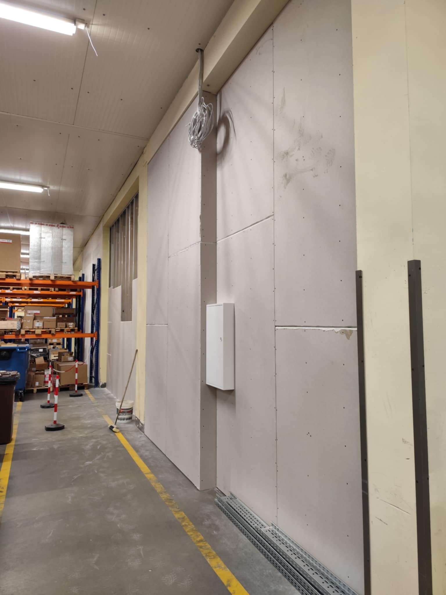 Wnętrze magazynu z ścianą z płyt karton-gips, widoczne kable i puszka elektryczna. Na podłodze żółta linia, w tle regały z towarem.
