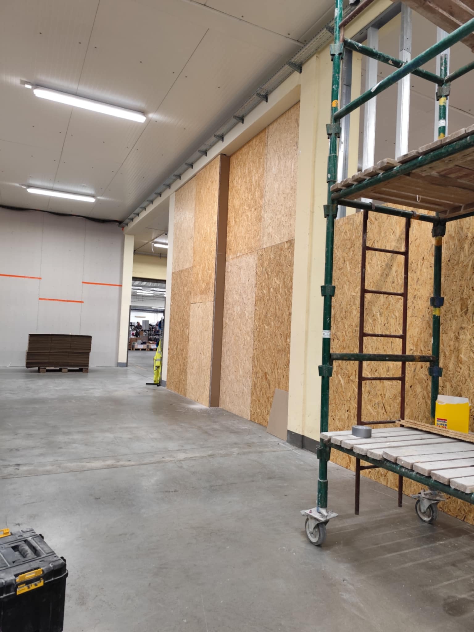 Wnętrze hali w trakcie prac wykończeniowych: obudowa ścian płytami OSB, widoczne rusztowanie, skrzynka narzędziowa i materiały budowlane.
