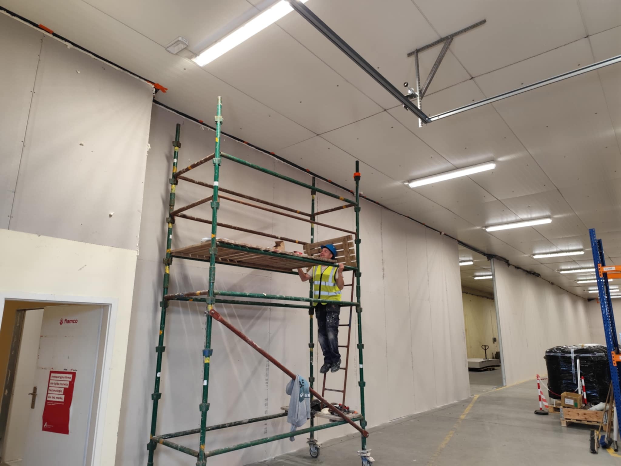 Pracownik na rusztowaniu montuje płyty gipsowo-kartonowe w hali. Widoczne ściany z płyt, oświetlenie sufitowe i elementy konstrukcyjne.