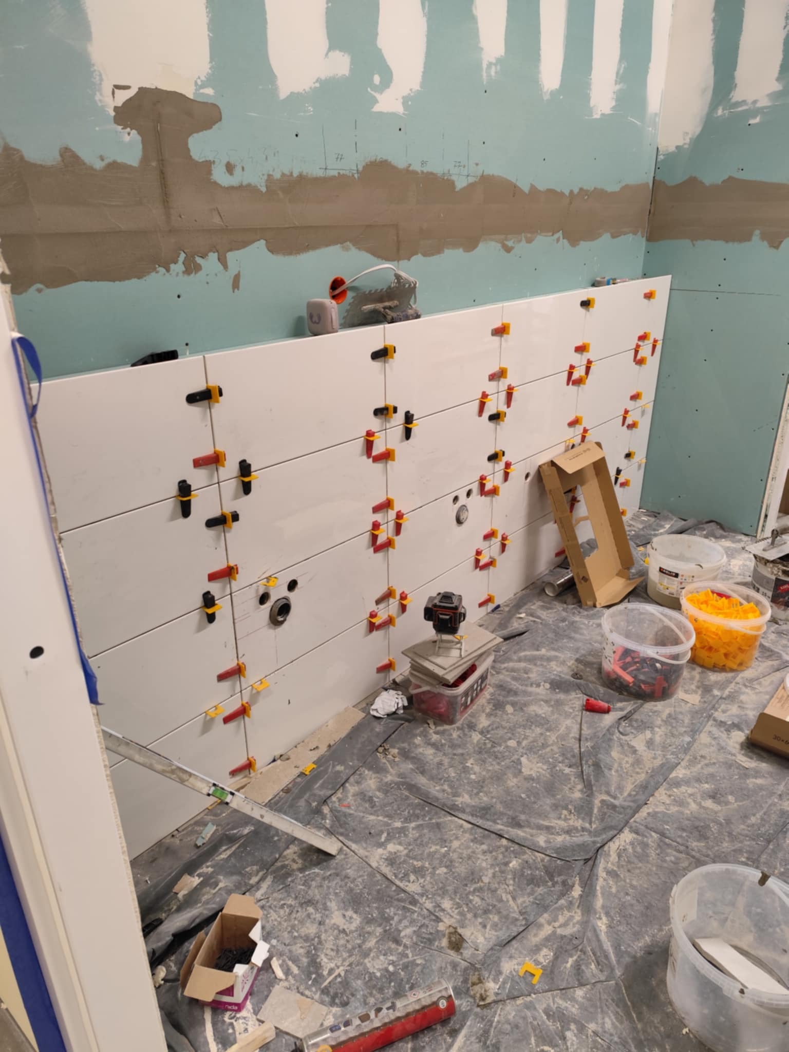 Ściana w trakcie układania białych, prostokątnych płytek, z użyciem kolorowych krzyżyków dystansowych. Widoczne narzędzia i materiały budowlane na podłodze.