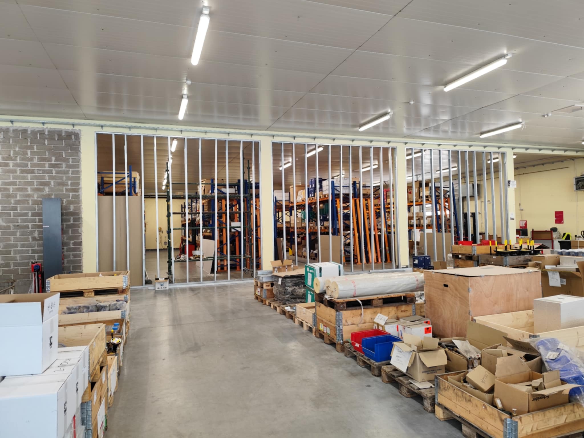 Magazyn w trakcie adaptacji: widoczne metalowe profile ścianek działowych, regały magazynowe w tle i materiały budowlane. Oświetlenie jarzeniowe, surowy stan wnętrza.