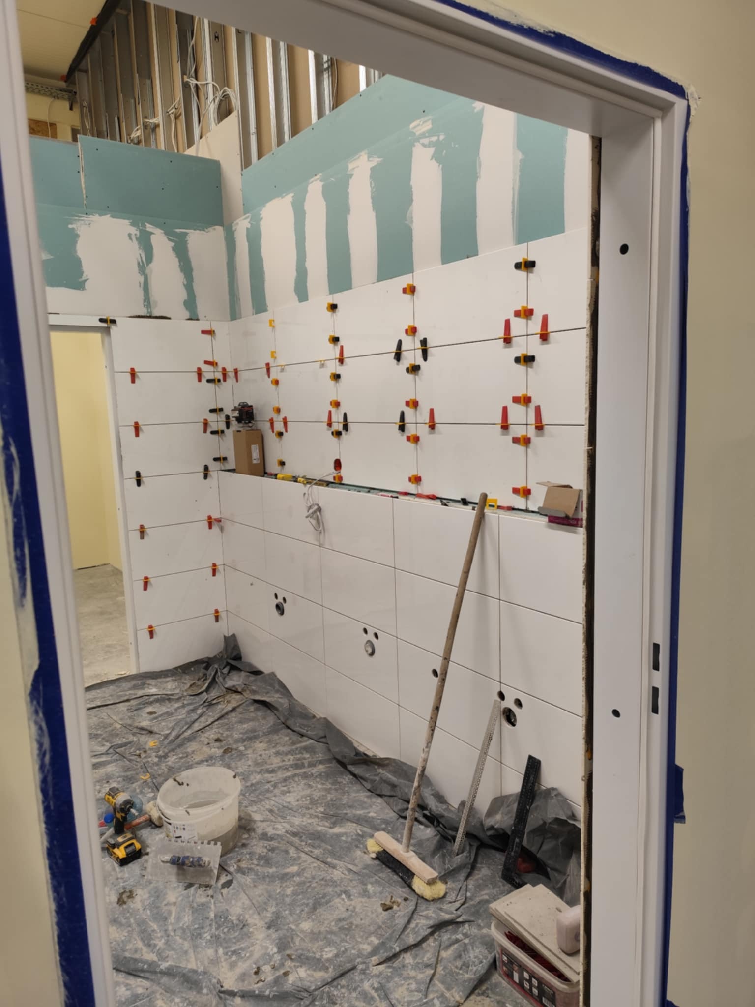 Białe płytki z krzyżykami dystansowymi na ścianie łazienki w trakcie remontu. Widoczne narzędzia glazurnicze i osłonięta folią podłoga. Ujęcie przez futrynę drzwi.