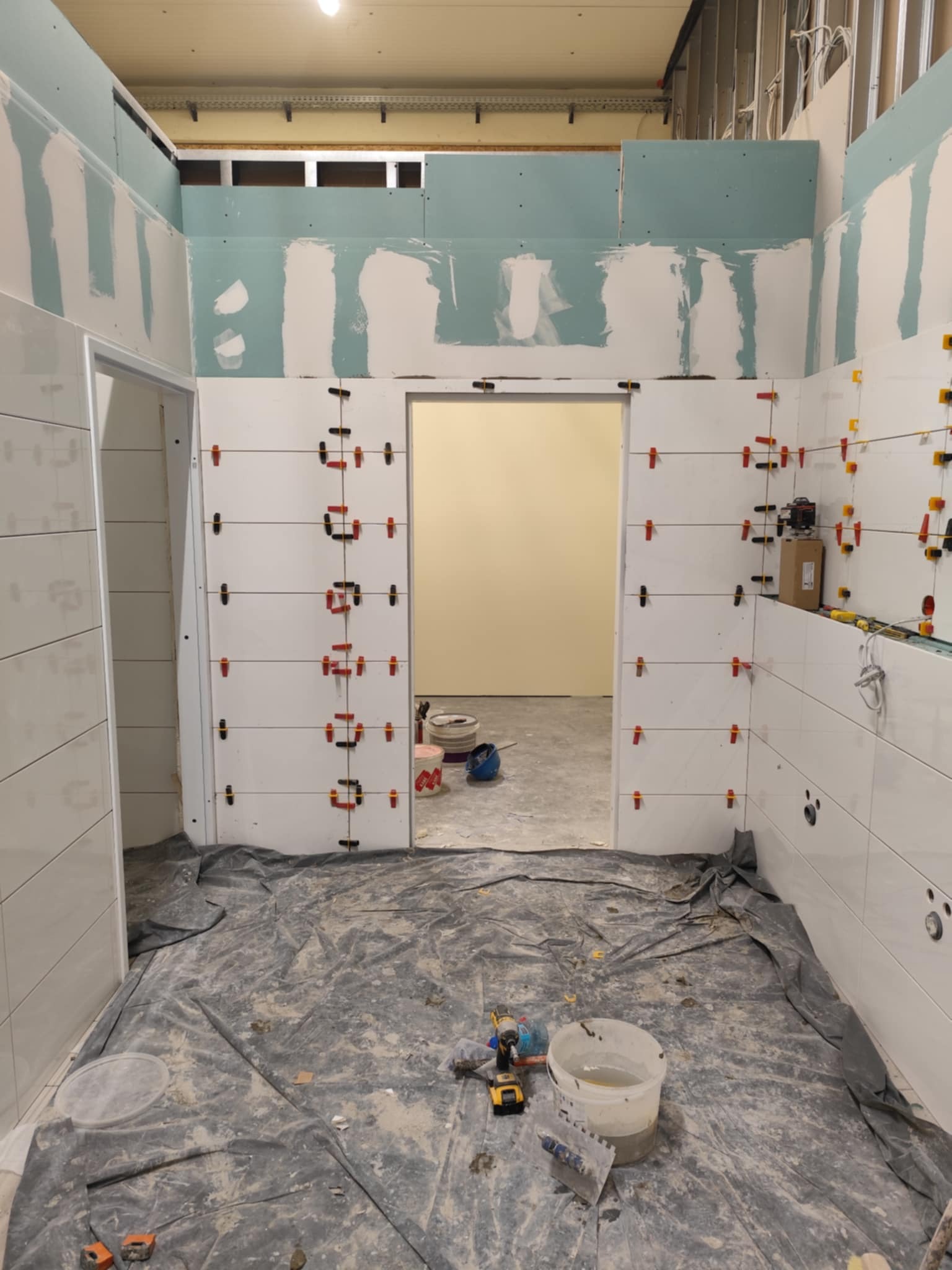 Białe płytki układane w łazience, z użyciem klipsów poziomujących. Widoczne narzędzia i folia ochronna na podłodze. Ściany częściowo wykończone płytami gipsowo-kartonowymi.