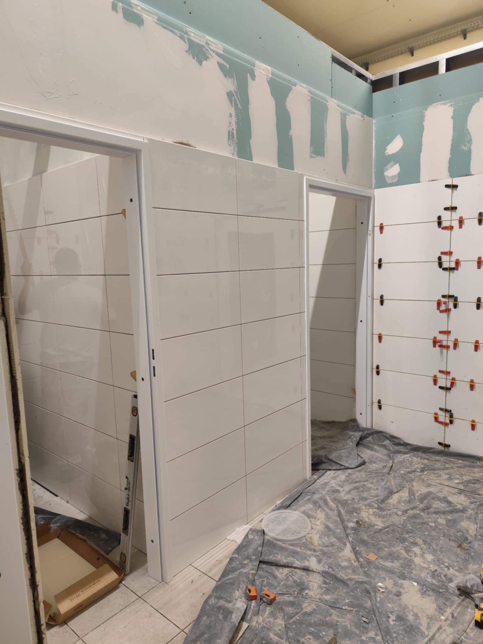 Remont łazienki w toku: białe, poziome płytki na ścianach, widoczne listwy wyrównujące, płyta karton gips na górze, zabezpieczona podłoga. Dwa otwory drzwiowe.