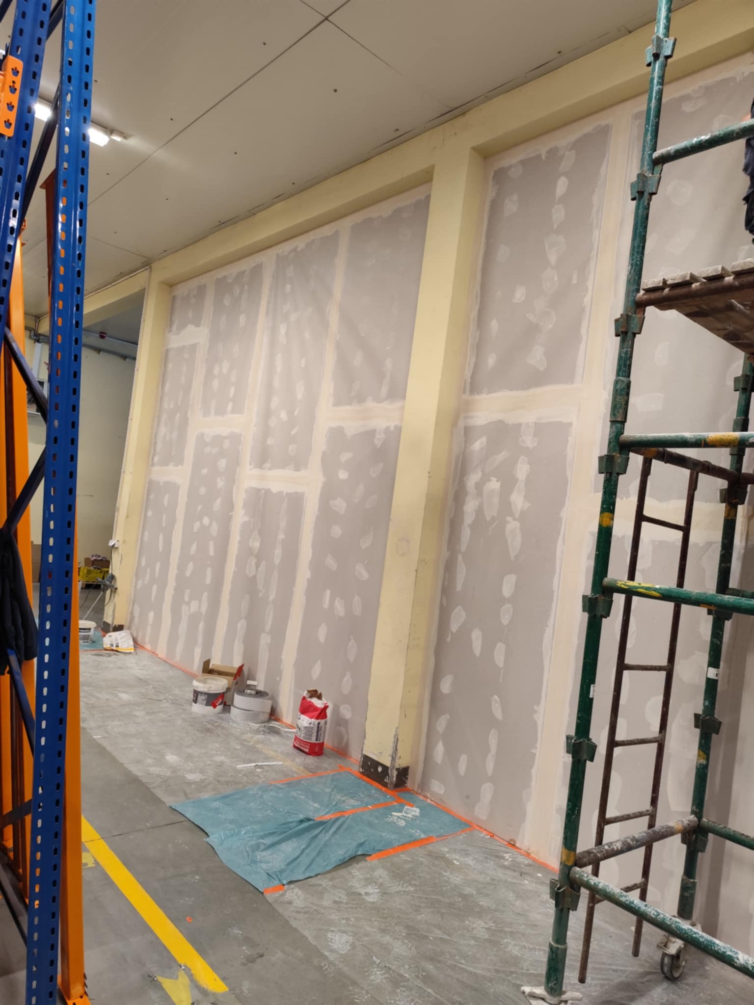 Ściana z płyt karton-gips w trakcie szpachlowania, widoczne rusztowanie i materiały budowlane. Żółte słupy konstrukcyjne, podłoga zabezpieczona folią.