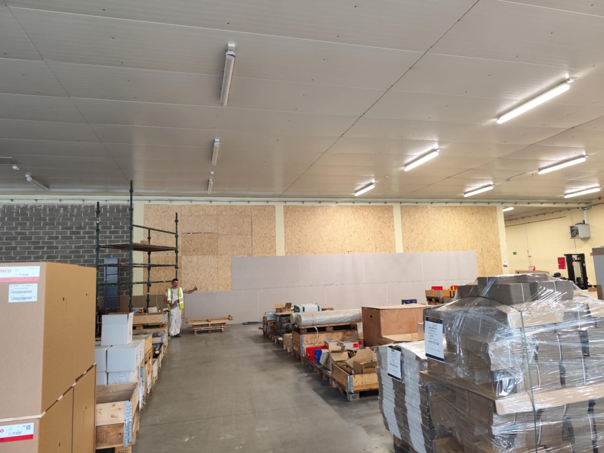 Wnętrze hali w trakcie prac wykończeniowych: ściany z płyt OSB, pracownik w odzieży roboczej, materiały budowlane na paletach. Widoczne oświetlenie sufitowe.