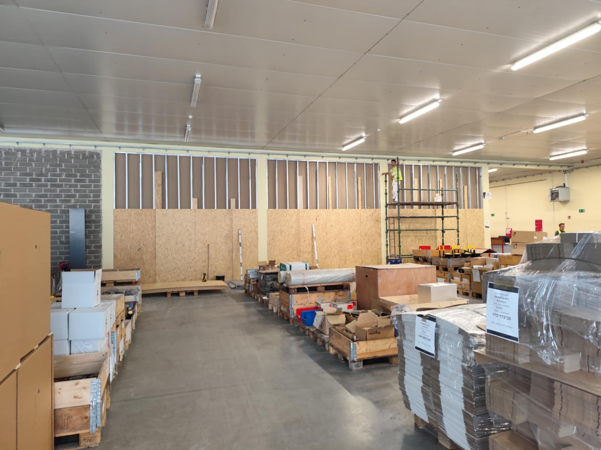 Duża przestrzeń magazynowa w trakcie prac wykończeniowych: montaż płyt OSB i konstrukcji stalowej. Pracownik na rusztowaniu. Materiały budowlane w paletach.