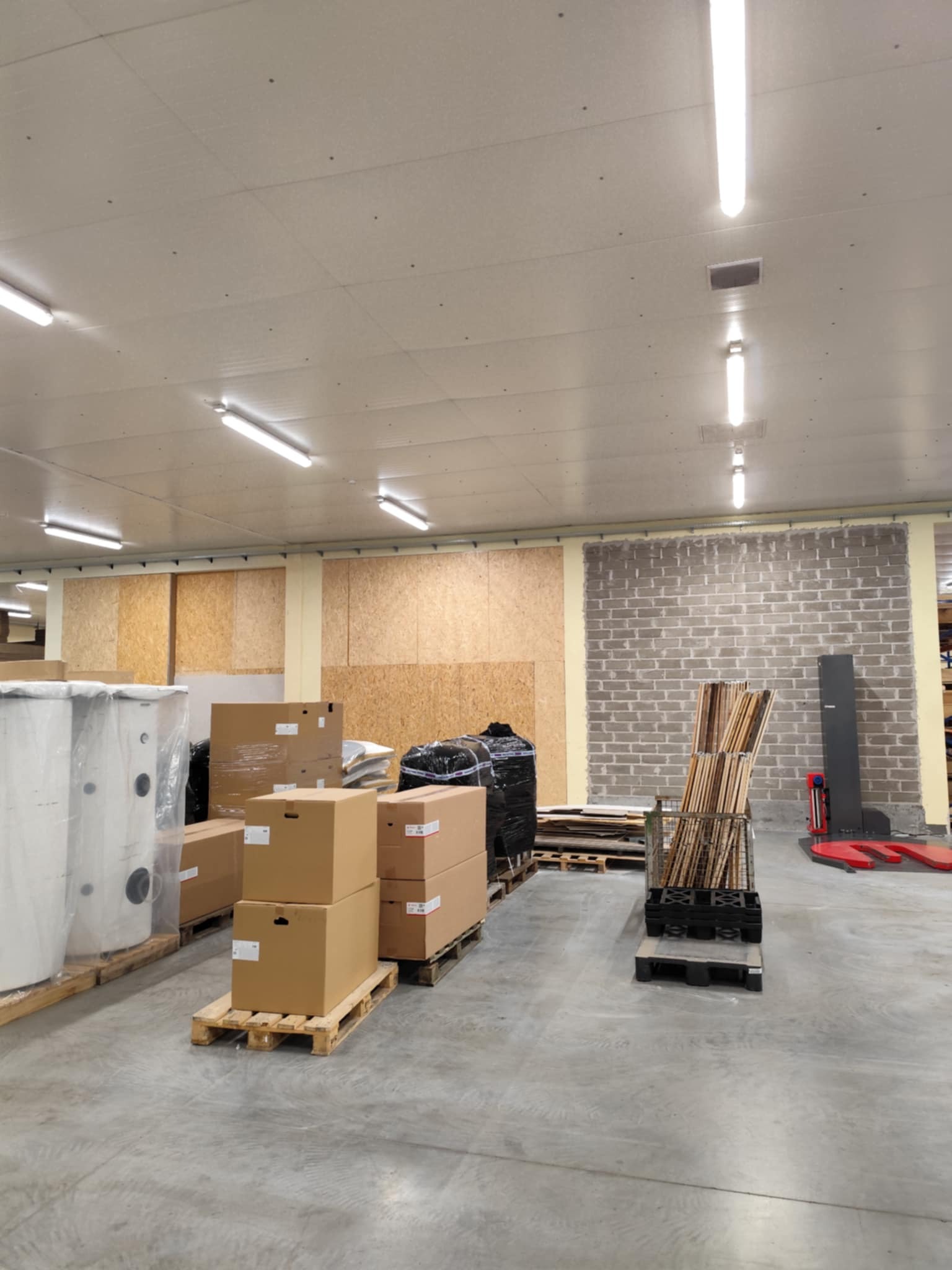 Magazyn z materiałami budowlanymi: kartony na paletach, płyty OSB i ceglana ściana. W tle widoczny wózek paletowy.