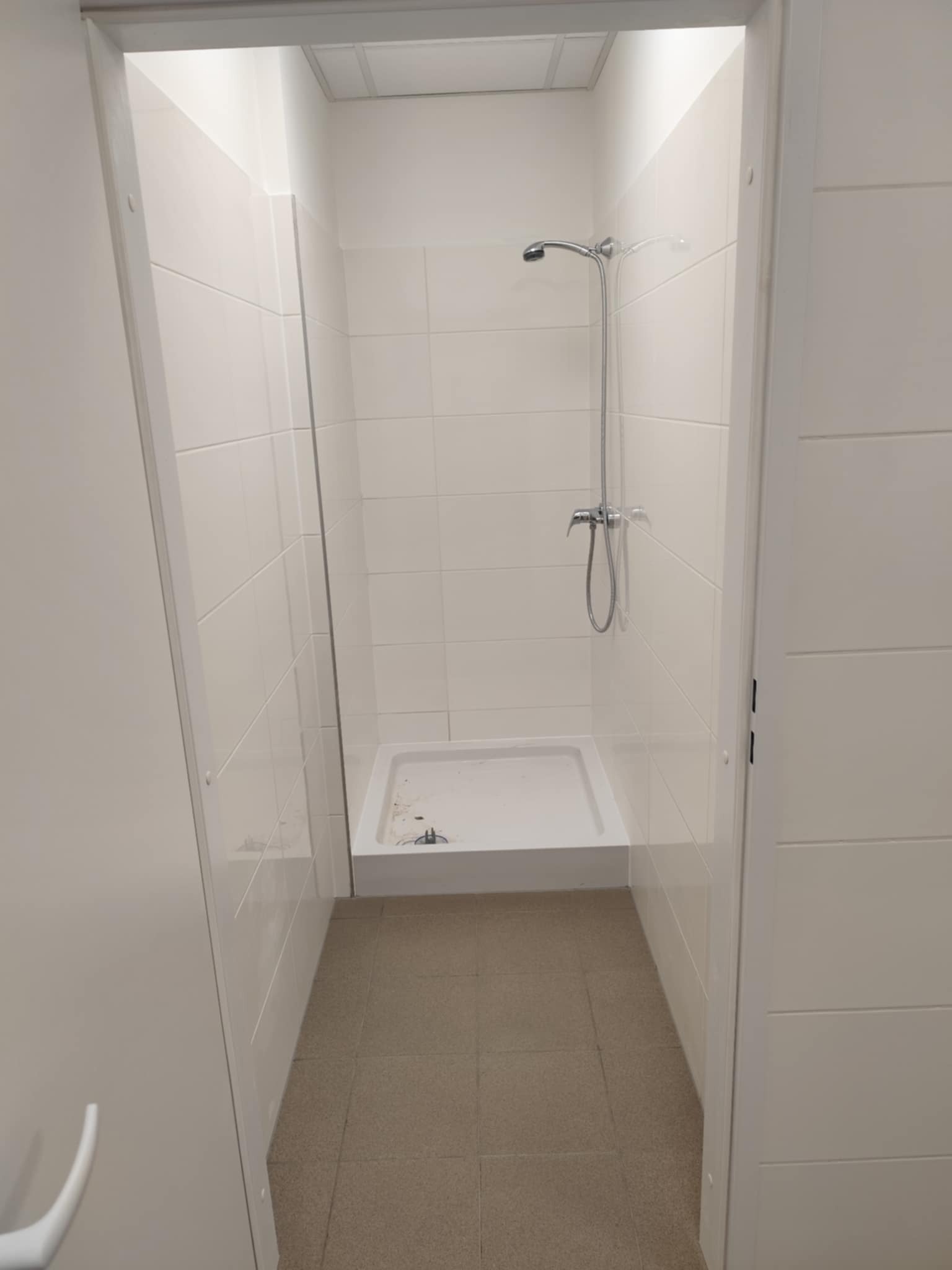 Wykończona kabina prysznicowa z białymi płytkami i brodzikiem, widoczna przez otwarte drzwi. Chromowana bateria i słuchawka prysznicowa. Podłoga wyłożona brązowymi płytkami.