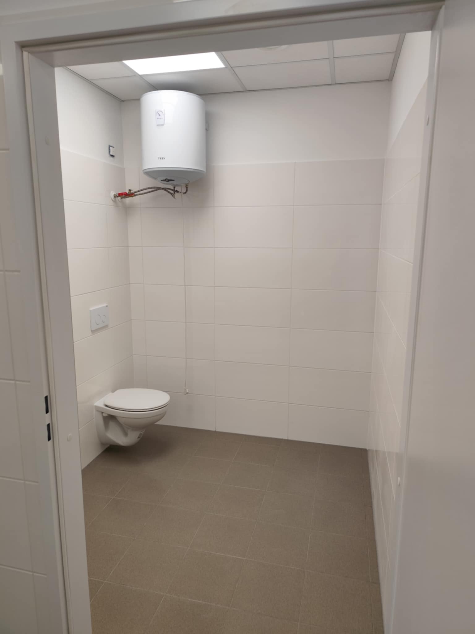 Jasna łazienka z białymi płytkami, toaletą i bojlerem. Brązowa podłoga z płytek. Widok przez otwarte drzwi. Minimalistyczny design.