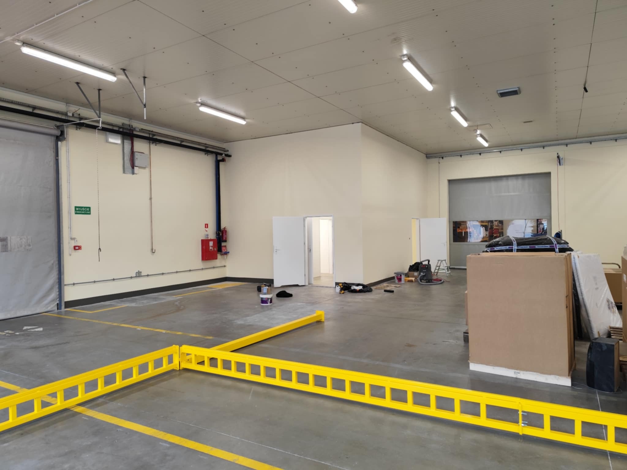 Duża hala magazynowa z pomalowanymi ścianami na beżowo, widoczne żółte linie na szarej posadzce. Świeżo po malowaniu.