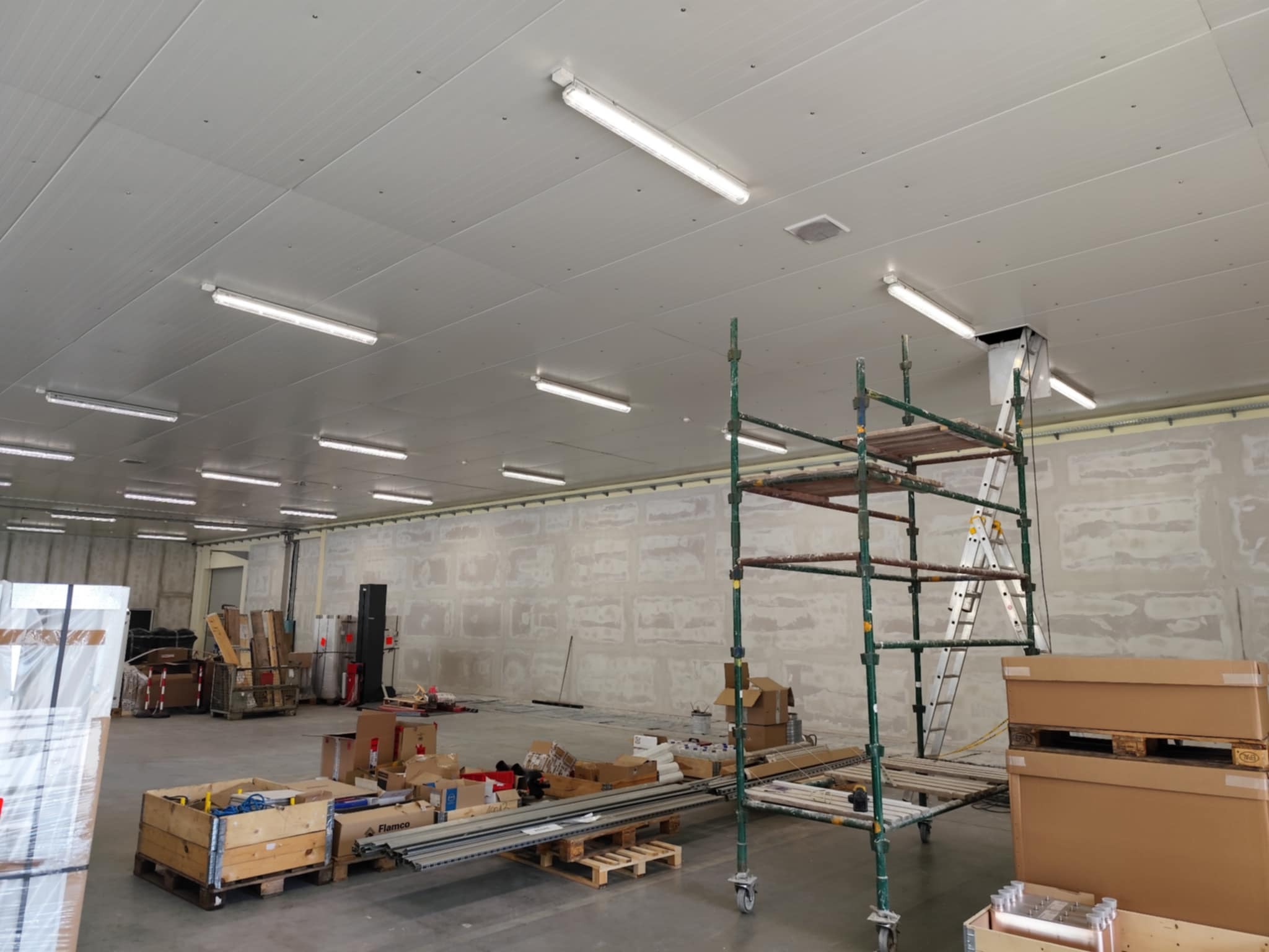 Wnętrze w trakcie wykończenia: sufit z oświetleniem, ściany przygotowane do malowania, rusztowanie, drabina i materiały budowlane na podłodze.