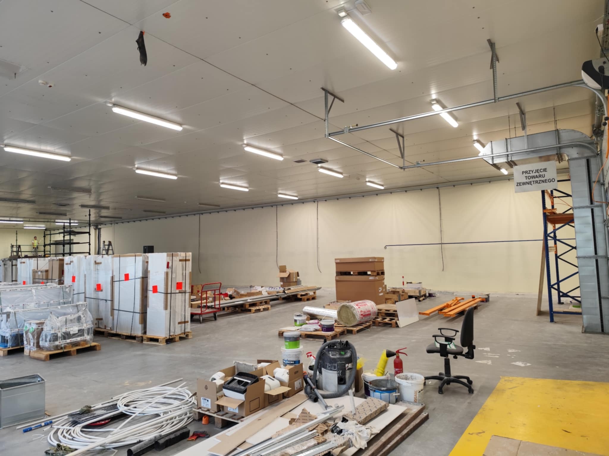 Wnętrze hali w trakcie prac wykończeniowych: materiały budowlane, farby, narzędzia, płyty. Widoczne instalacje pod sufitem i regały magazynowe.