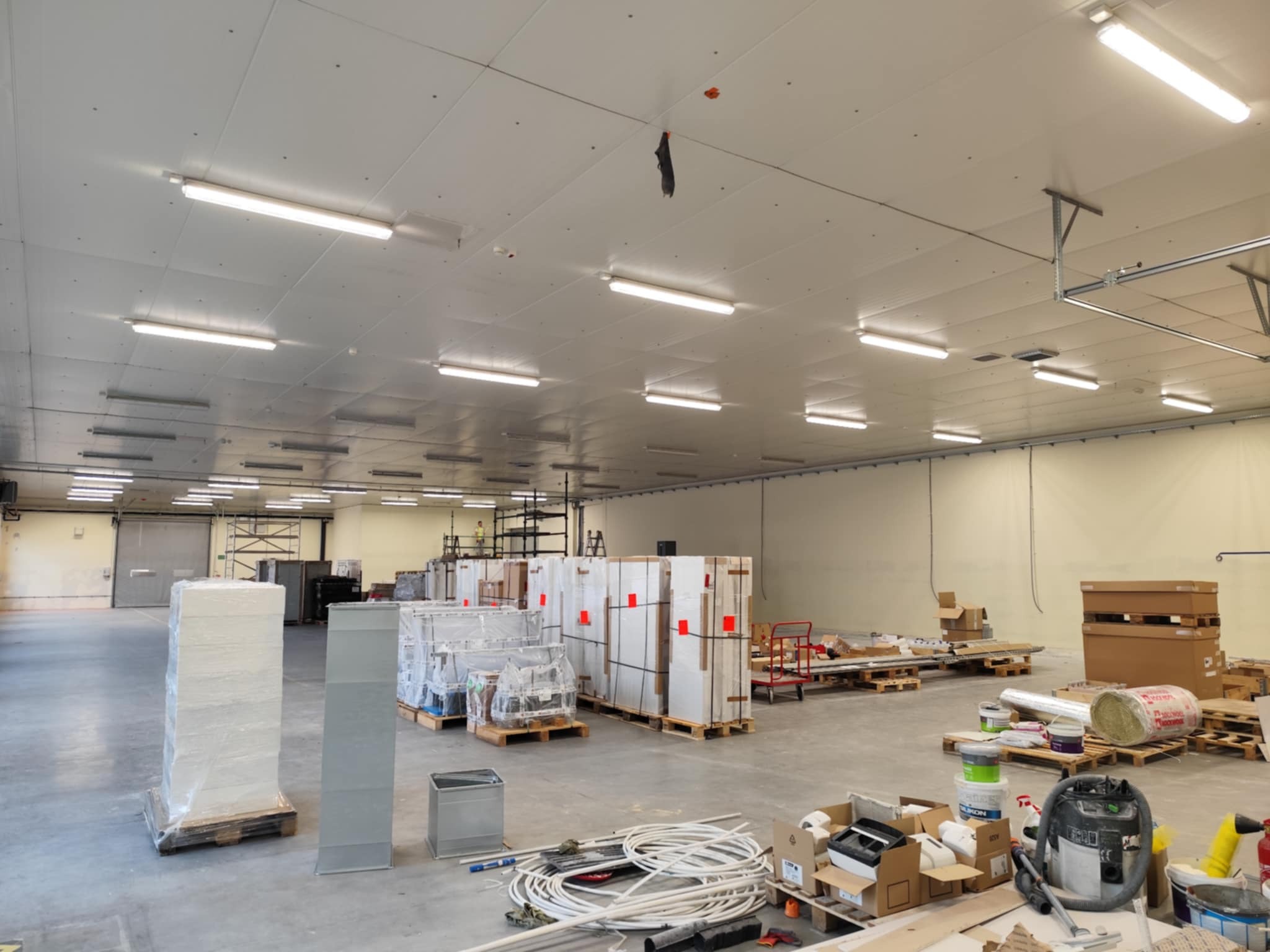 Wnętrze hali w trakcie prac wykończeniowych. Widoczne materiały budowlane, narzędzia, oświetlenie ledowe pod sufitem i elementy wentylacji. Część materiałów zabezpieczona folią.