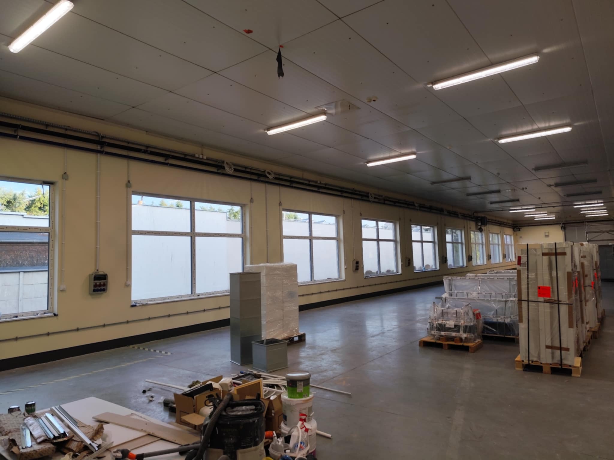 Wnętrze hali w trakcie prac wykończeniowych: okna z białymi ramami, materiały budowlane na podłodze, przygotowane do montażu elementy. Oświetlenie jarzeniowe.