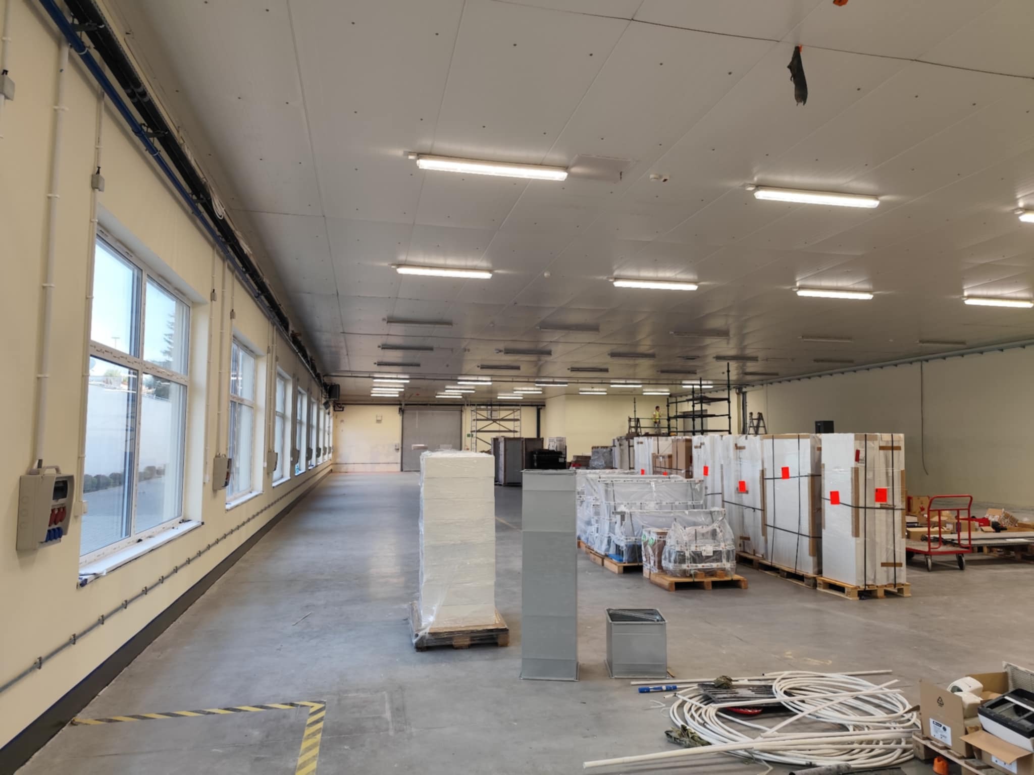 Przestronne wnętrze hali produkcyjnej z rzędem okien wzdłuż ściany, widocznymi instalacjami elektrycznymi i wentylacyjnymi oraz materiałami budowlanymi przygotowanymi do montażu.