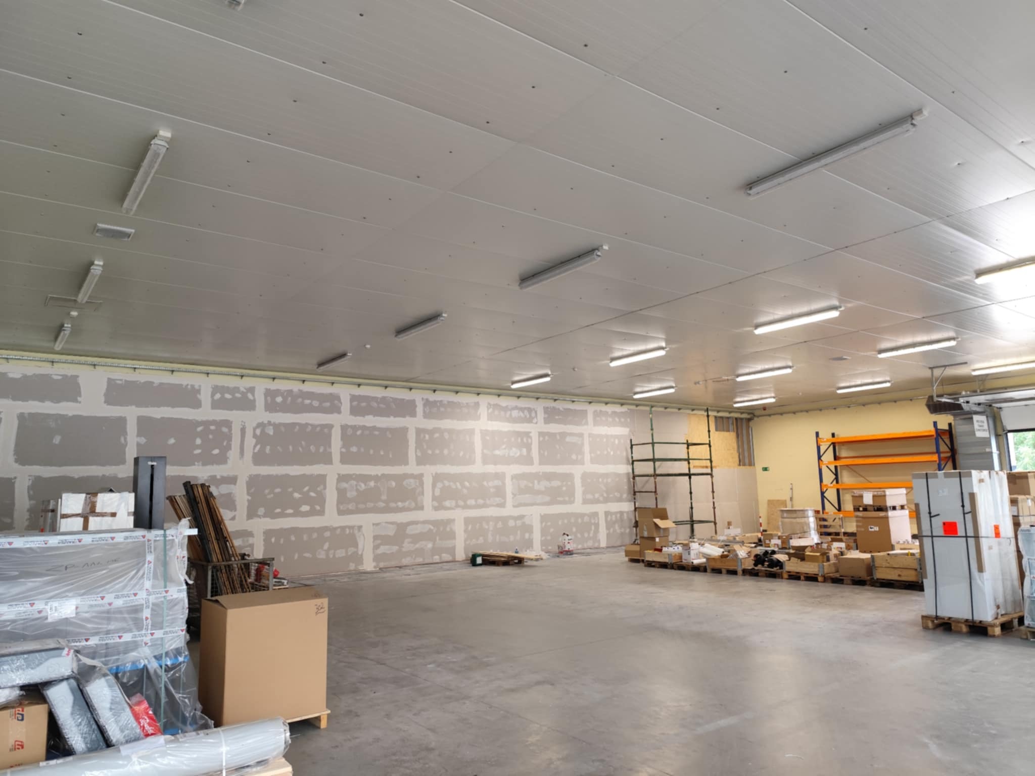 Wnętrze hali w trakcie prac wykończeniowych. Ściany z widocznymi płytami kartonowo-gipsowymi przed szpachlowaniem. Sufit z oświetleniem rastrowym.