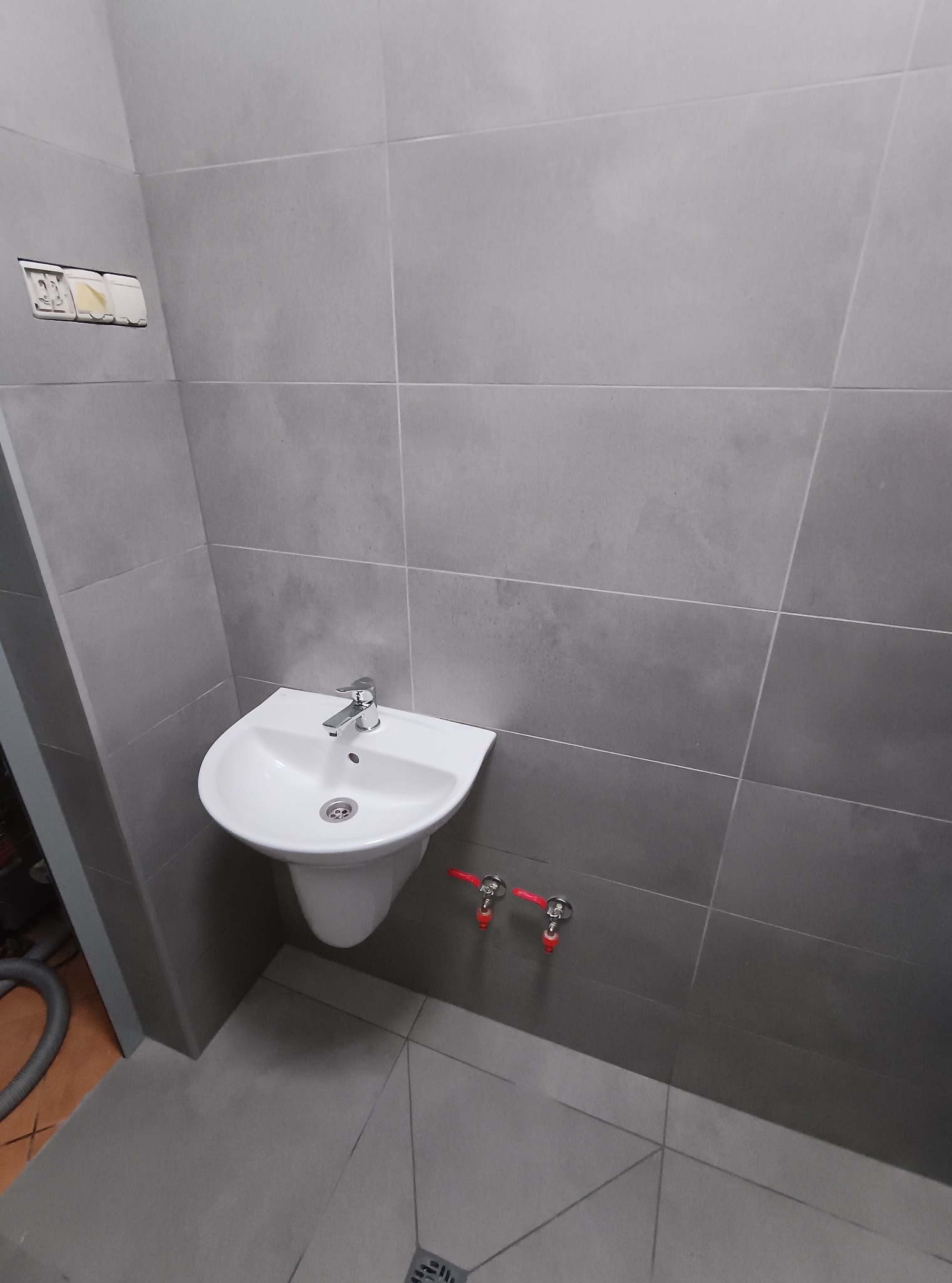 Szara łazienka po remoncie: biała umywalka z chromowaną baterią na tle płytek. Widoczne zawory z czerwonymi kurkami i fragment odkurzacza. Gniazdko elektryczne bez osprzętu.