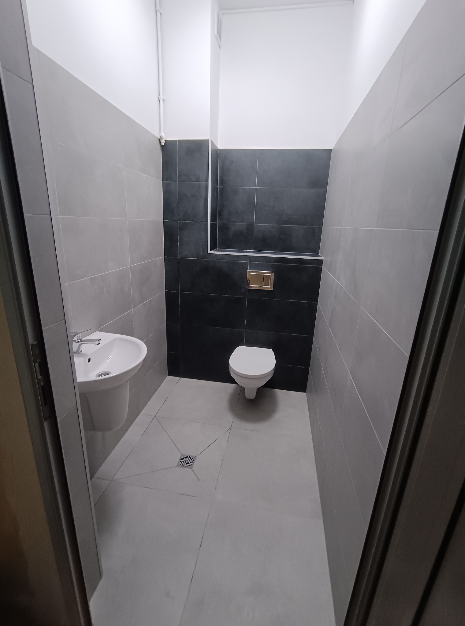 Nowoczesna łazienka po remoncie: szare i czarne płytki, biała umywalka i toaleta. Widok zza otwartych drzwi, minimalistyczny design, czysta przestrzeń.