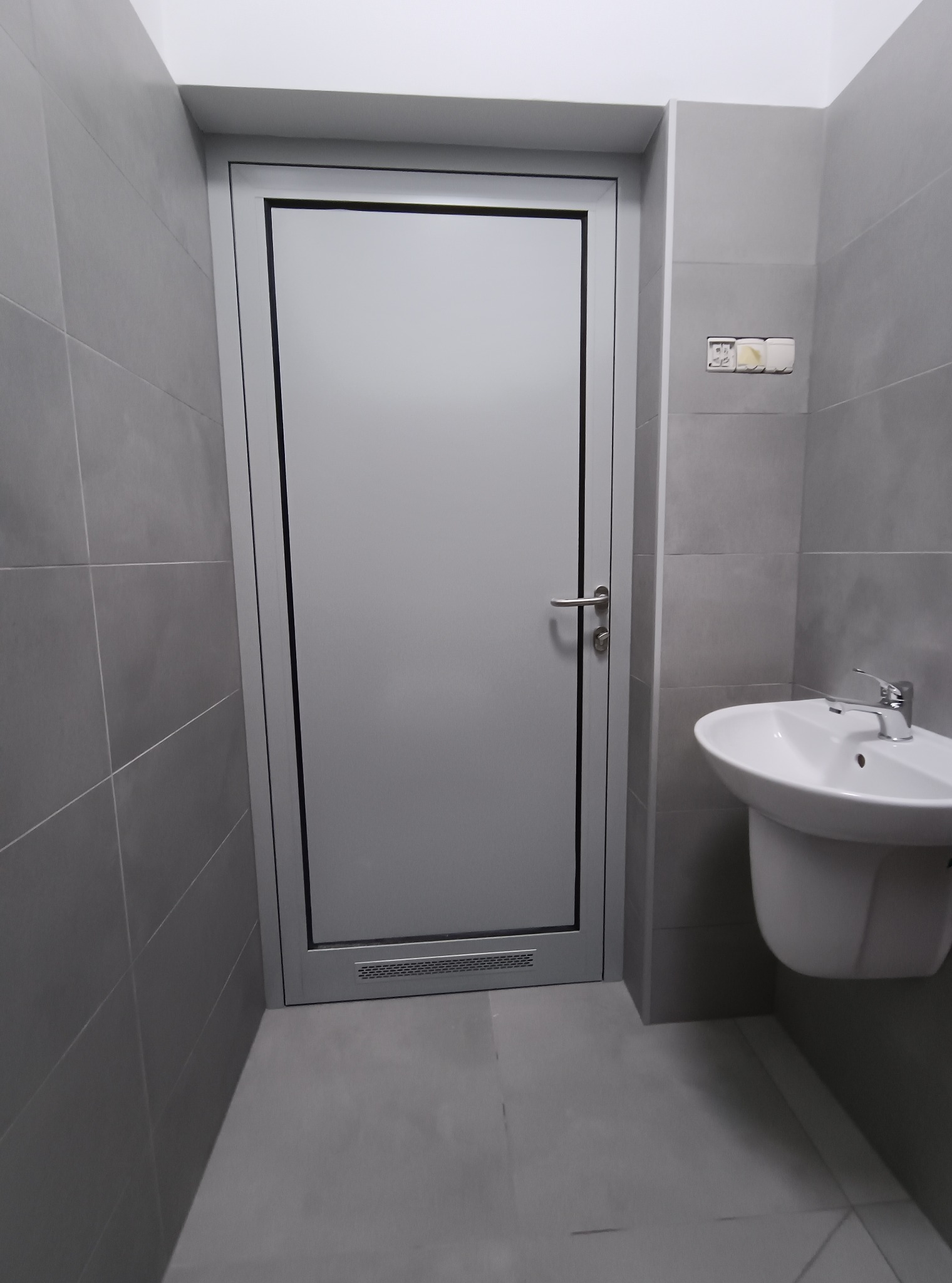 Szara łazienka z umywalką i drzwiami. Płytki na ścianach i podłodze w odcieniach szarości. Minimalistyczny design, widoczne gniazdka elektryczne.