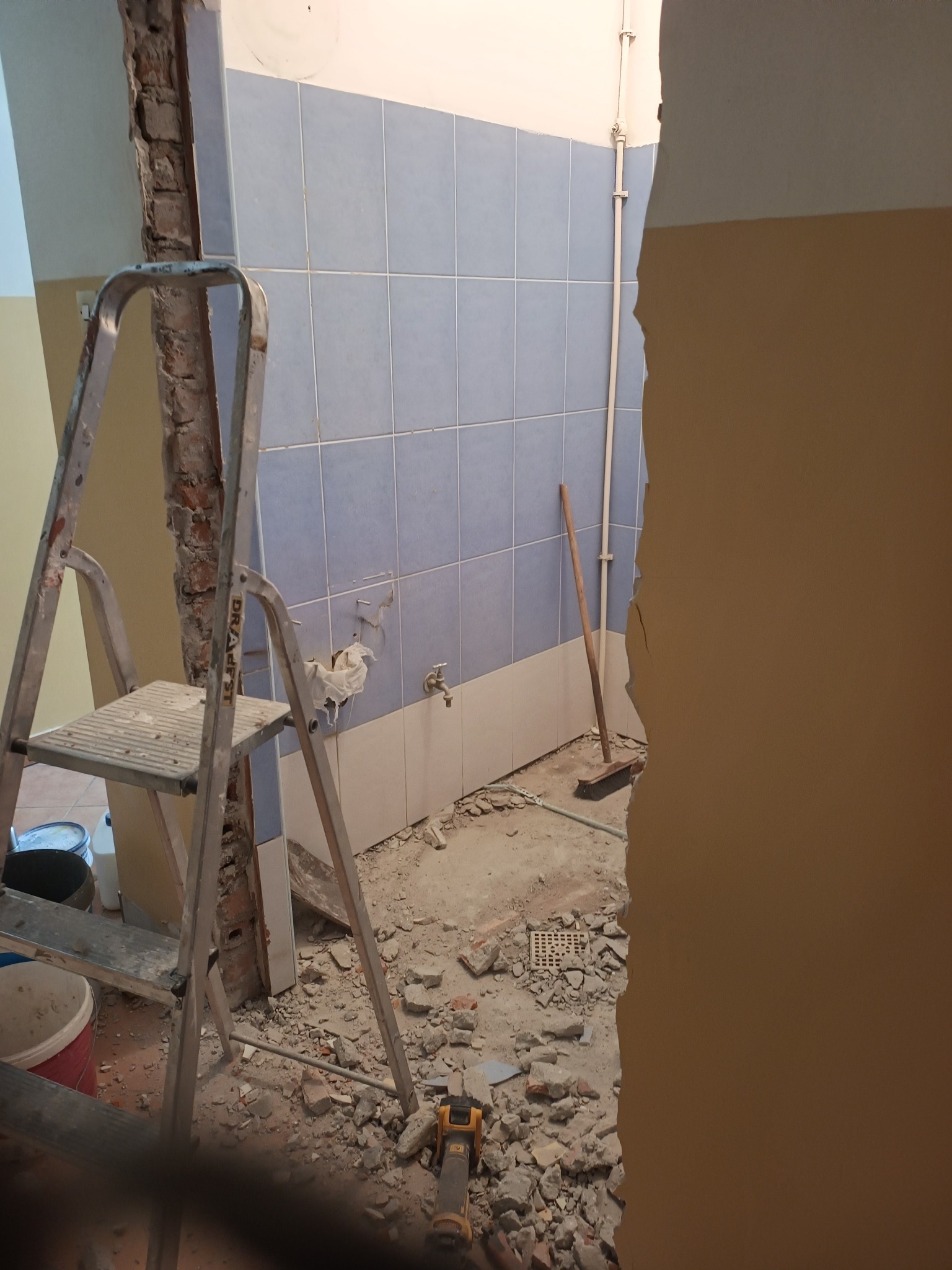 Demontaż płytek w łazience podczas remontu. Widoczna drabina, gruz i fragment ściany z niebieskimi płytkami. Częściowo usunięte płytki odsłaniają instalację.
