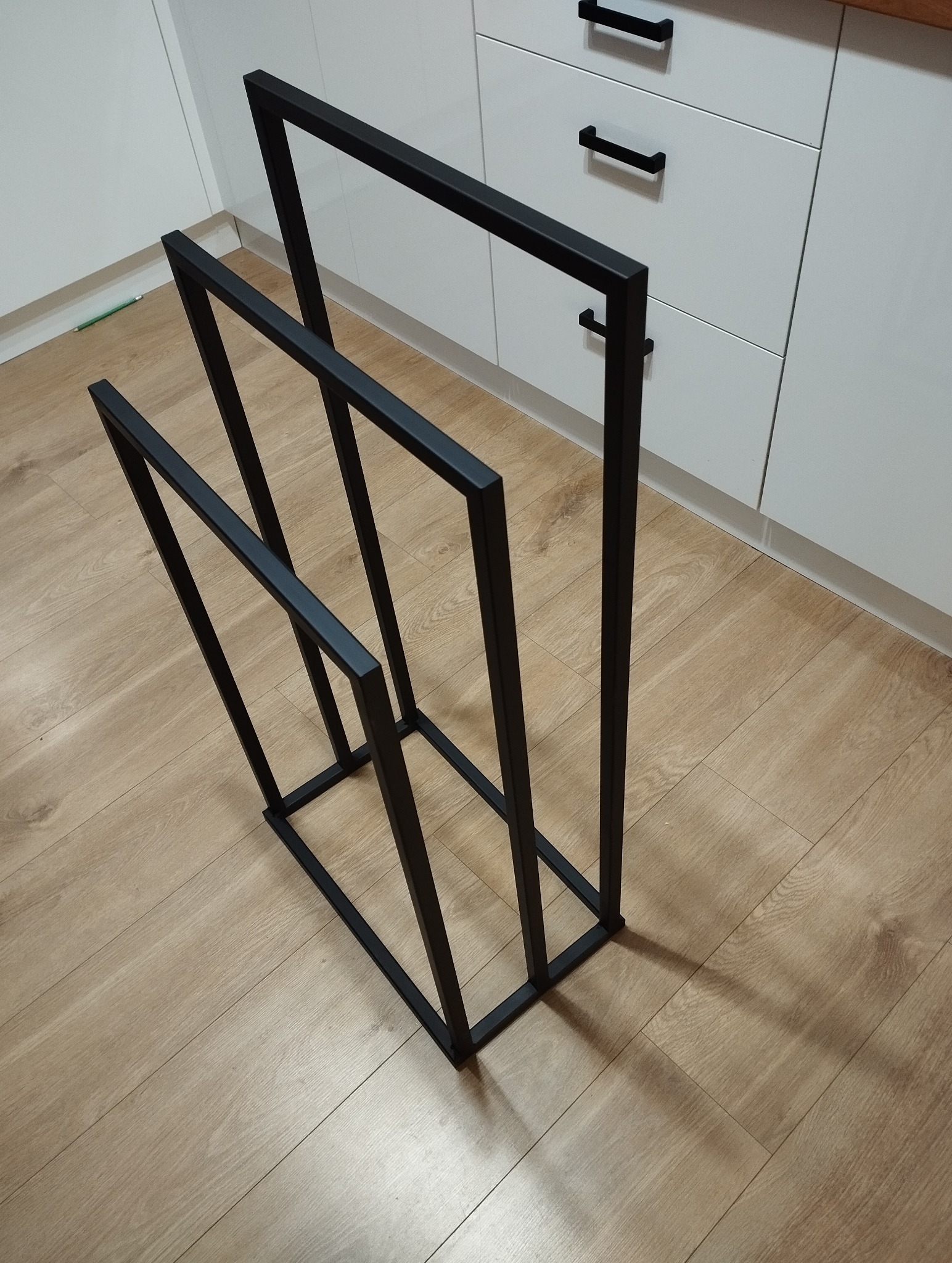 Czarny, minimalistyczny stojak na ręczniki z metalowych profili, ustawiony na podłodze z paneli, na tle białych szafek kuchennych z czarnymi uchwytami.