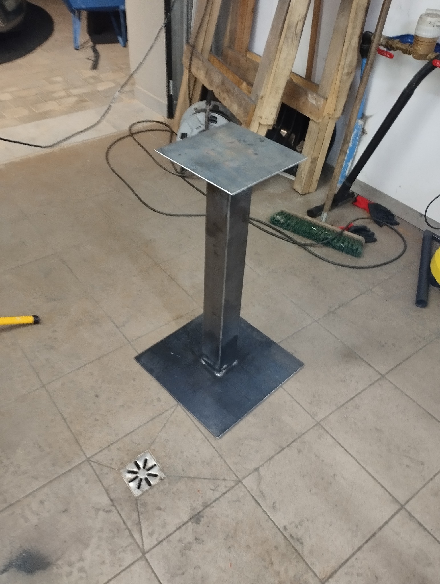 Surowa, spawana konstrukcja metalowa z kwadratową podstawą i blatem, stojąca na kafelkach w warsztacie. Widoczne narzędzia i akcesoria w tle.