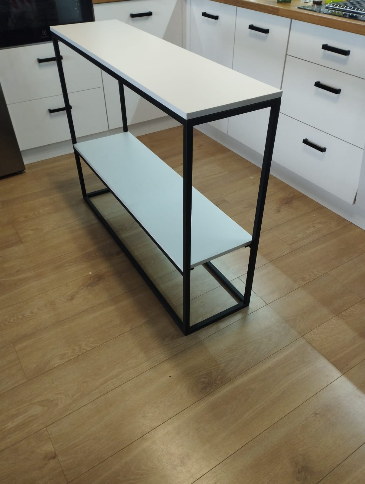 Metalowa konstrukcja z dwoma białymi blatami, czarne profile, minimalistyczny design, na tle białych szafek kuchennych i drewnianej podłogi.