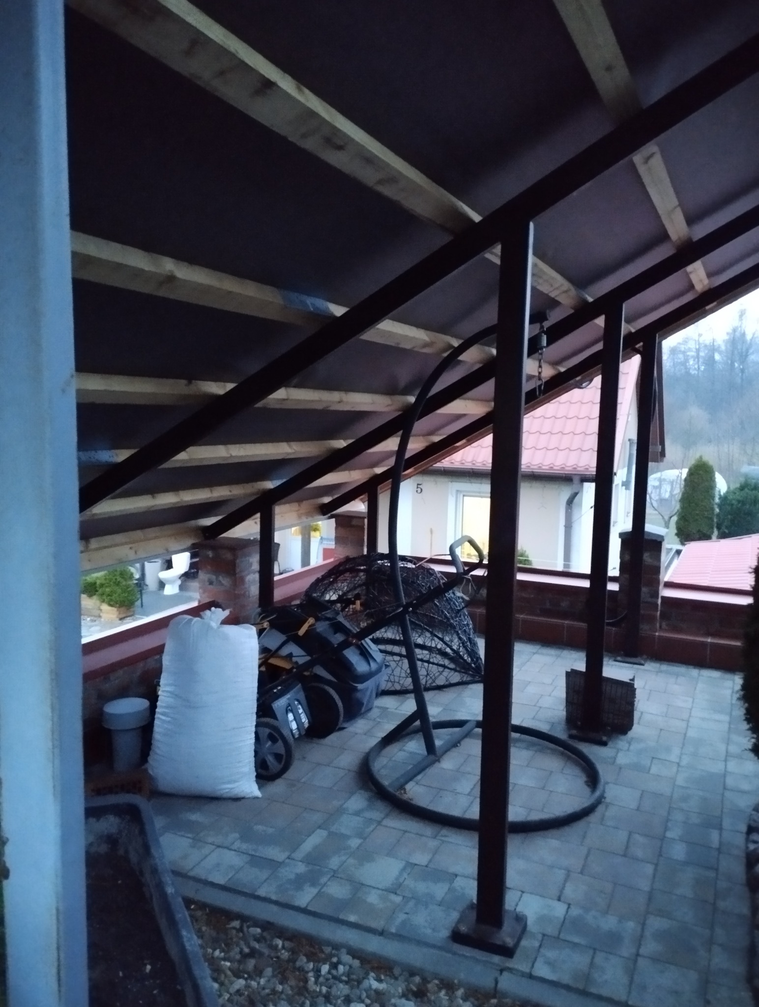 Zadaszenie tarasu z metalową konstrukcją, widok z wnętrza. Na tarasie worek z materiałami budowlanymi i huśtawka typu kokon. W tle dom z czerwonym dachem.