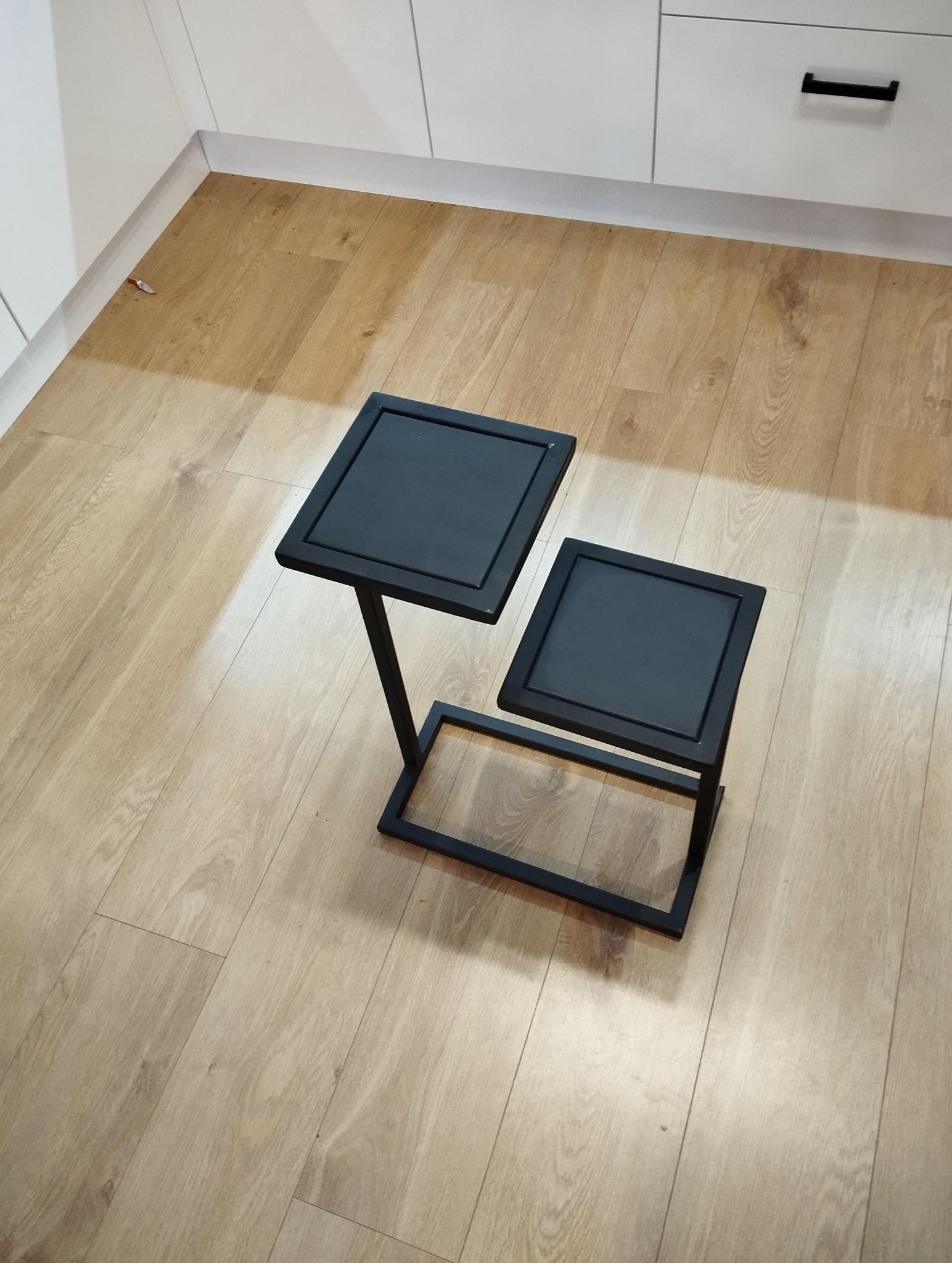 Dwa czarne, metalowe stoliki kawowe o kwadratowych blatach, ustawione na drewnianej podłodze w jasnym wnętrzu. Minimalistyczny design, widoczne białe szafki w tle.