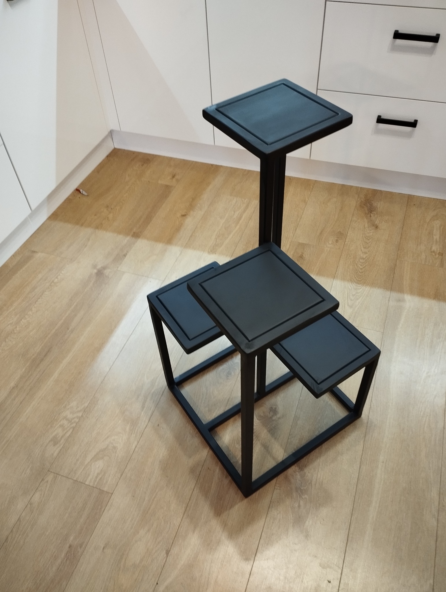 Czarny, metalowy kwietnik trójpoziomowy w minimalistycznym stylu, na tle białych szafek kuchennych i drewnianej podłogi. Geometryczna konstrukcja z kwadratowymi blatami.