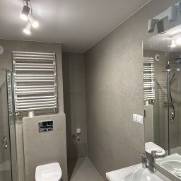 Nowoczesna łazienka w szarościach z białym wyposażeniem: toaleta, umywalka z szafką, grzejnik drabinkowy i kabina prysznicowa. Minimalistyczny design, oświetlenie punktowe.