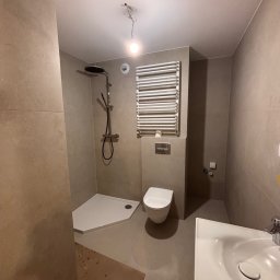 Wykończona łazienka w beżowych tonacjach z kabiną prysznicową, toaletą i grzejnikiem drabinkowym. Widoczne płytki na ścianach i podłodze, minimalistyczny design.