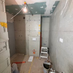 Wnętrze łazienki w trakcie remontu: szare płytki na ścianach, widoczne krzyżyki dystansowe, przygotowane podłoże sufitu, drabina, narzędzia i materiały budowlane.