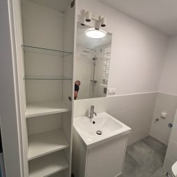 ANATOLII PRYIMAK BYGG - Biała, minimalistyczna łazienka po remoncie: szafka z umywalką, lustro z oświetleniem i wysoka szafka z półkami. Szare płytki podłogowe. Widoczne odbicie osoby w lustrze.