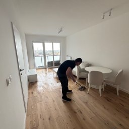 ANATOLII PRYIMAK BYGG - Mężczyzna myje drewnianą podłogę w jasnym, minimalistycznym wnętrzu z balkonem i widokiem na miasto. Widoczne ślady wilgoci po myciu.