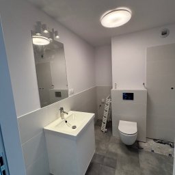 ANATOLII PRYIMAK BYGG - Nowoczesna, biała łazienka po remoncie z umywalką, lustrem i toaletą. Widoczne szare płytki podłogowe i białe ściany. Minimalistyczny design z oświetleniem sufitowym.