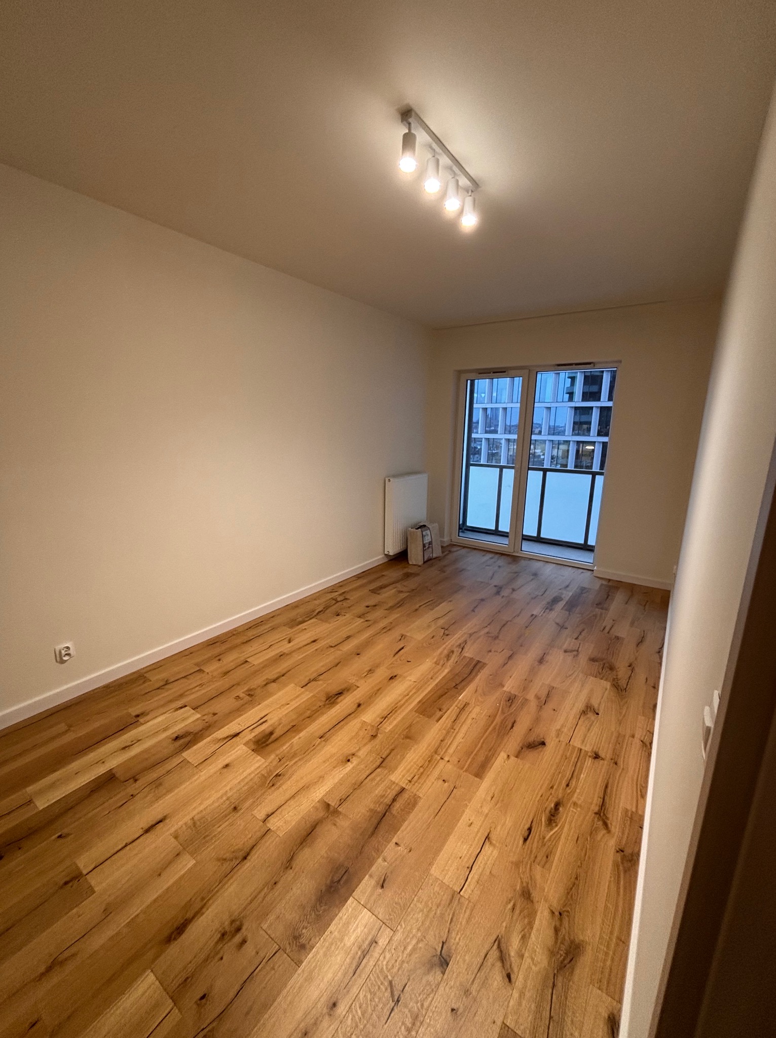 Jasne panele podłogowe w pustym pokoju z balkonem i grzejnikiem. Widoczne gniazdko elektryczne i listwa sufitowa. Oświetlenie sufitowe punktowe.