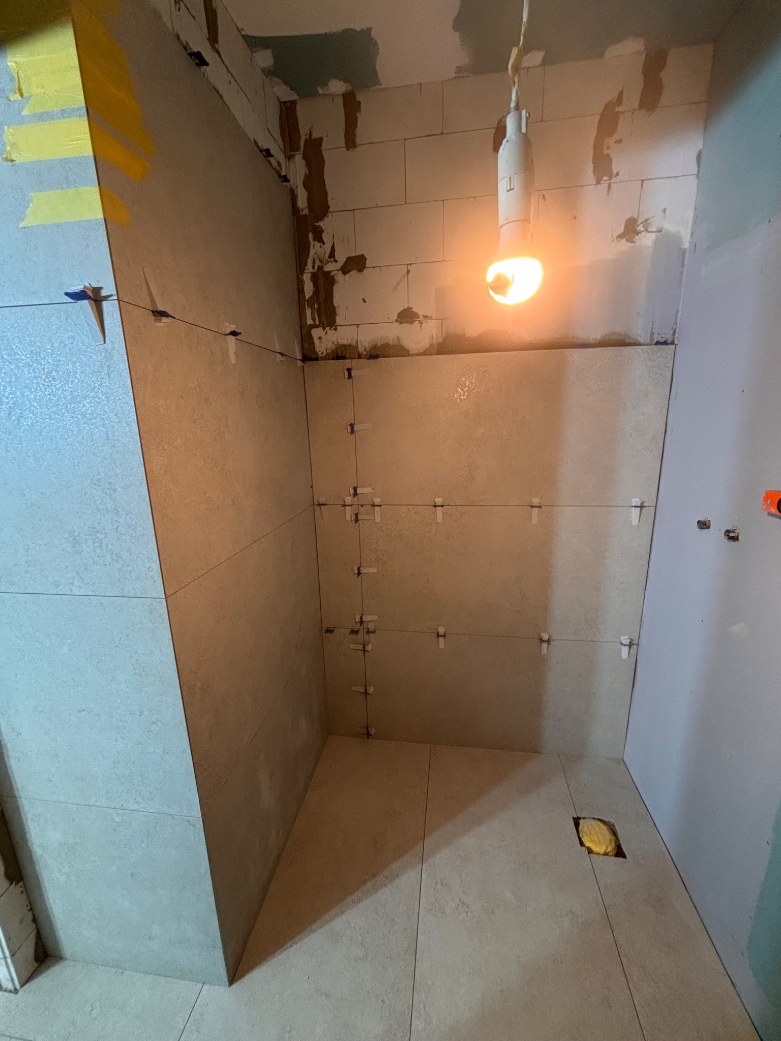 Wnętrze łazienki w trakcie prac wykończeniowych: ułożone płytki ścienne i podłogowe, widoczne krzyżyki dystansowe, surowy stan ścian w górnej części. Żarówka oświetla przestrzeń.