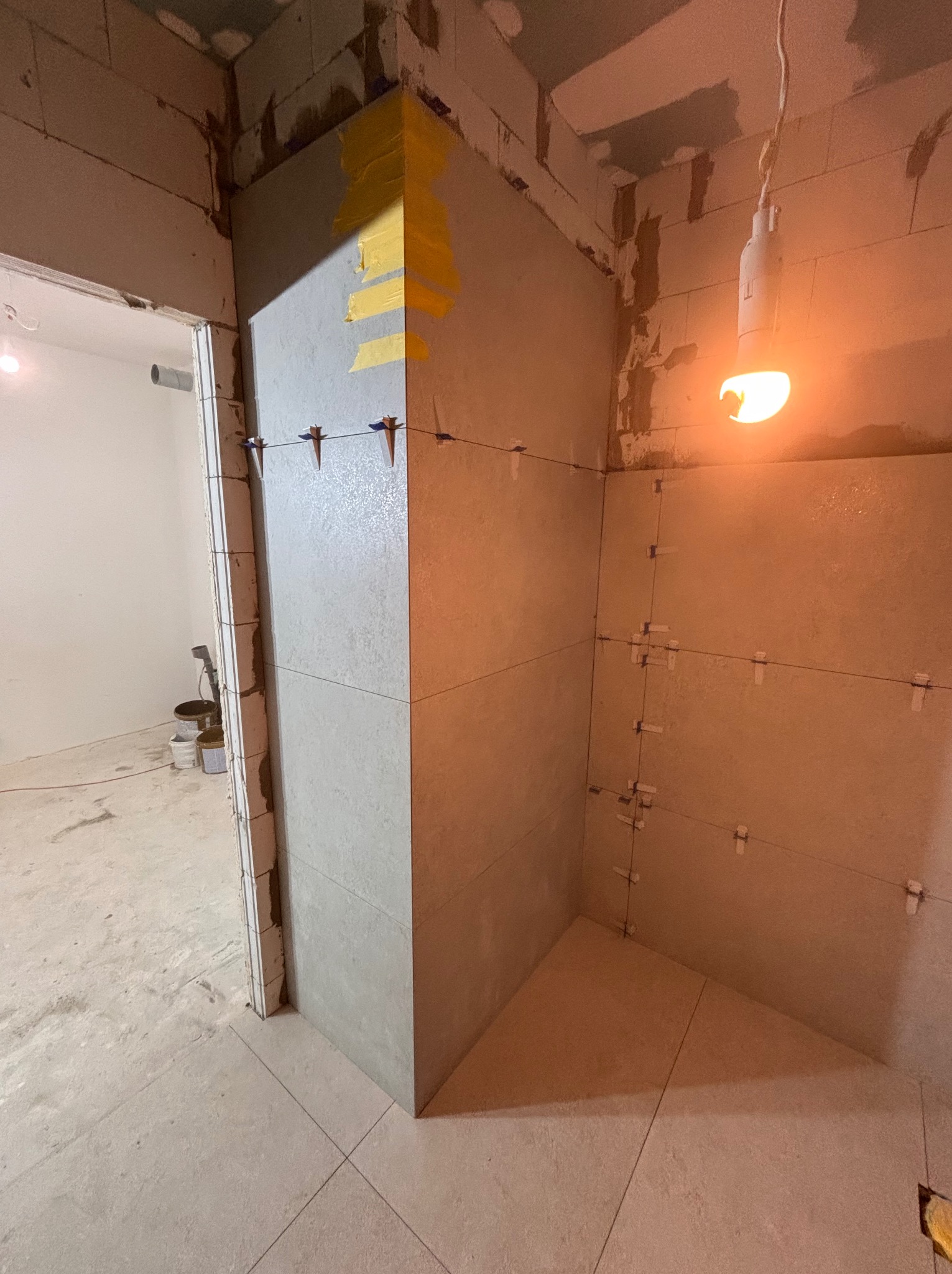 Ściana w trakcie układania płytek, widoczne krzyżyki dystansowe i taśma malarska na narożniku. Surowy stan pomieszczenia, wisząca żarówka.
