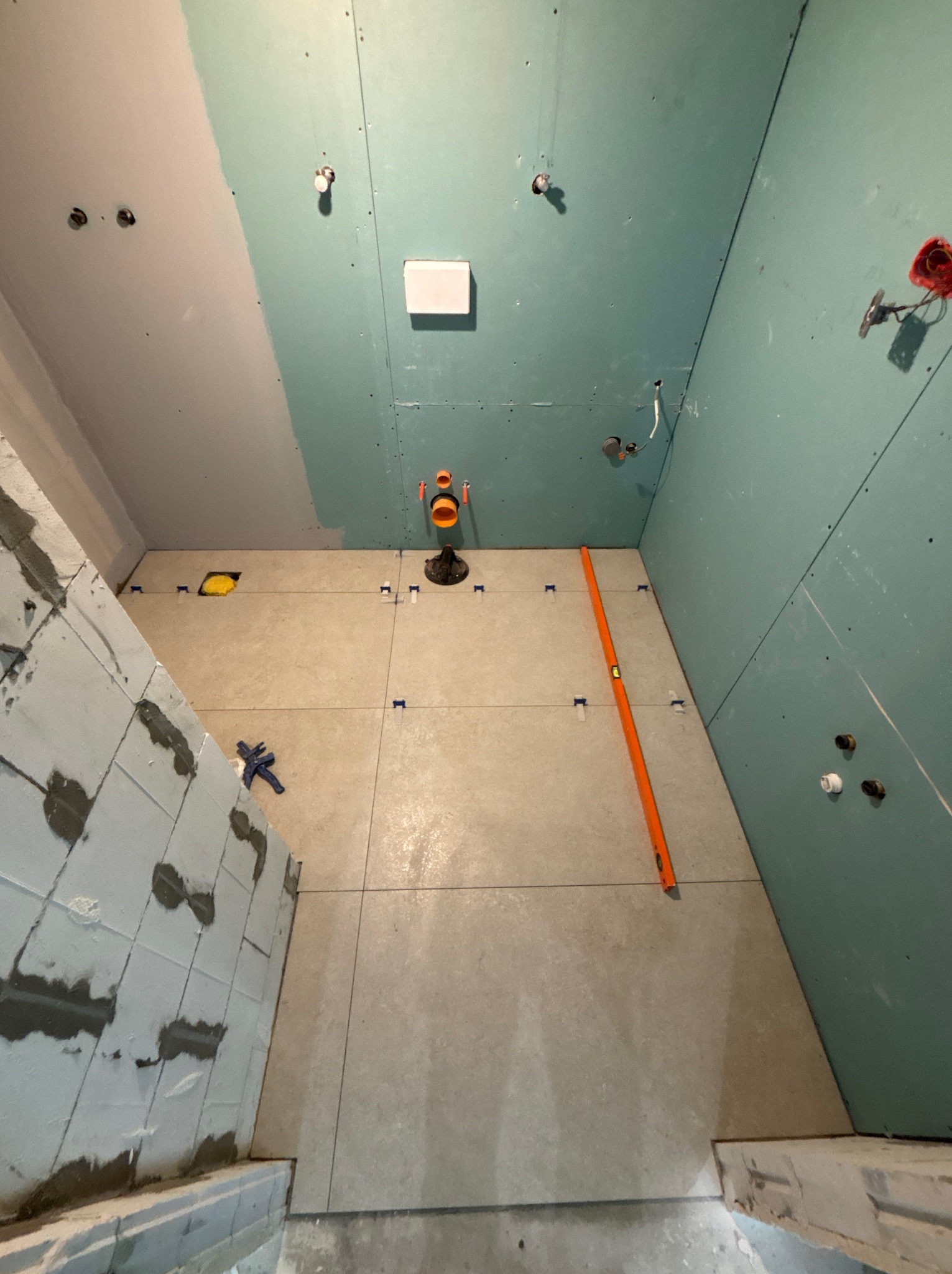 Wnętrze łazienki w trakcie prac: ułożone płytki podłogowe, częściowo wykończona ściana z płyty karton-gips, poziomica oparta o ścianę. Widoczne elementy instalacji.