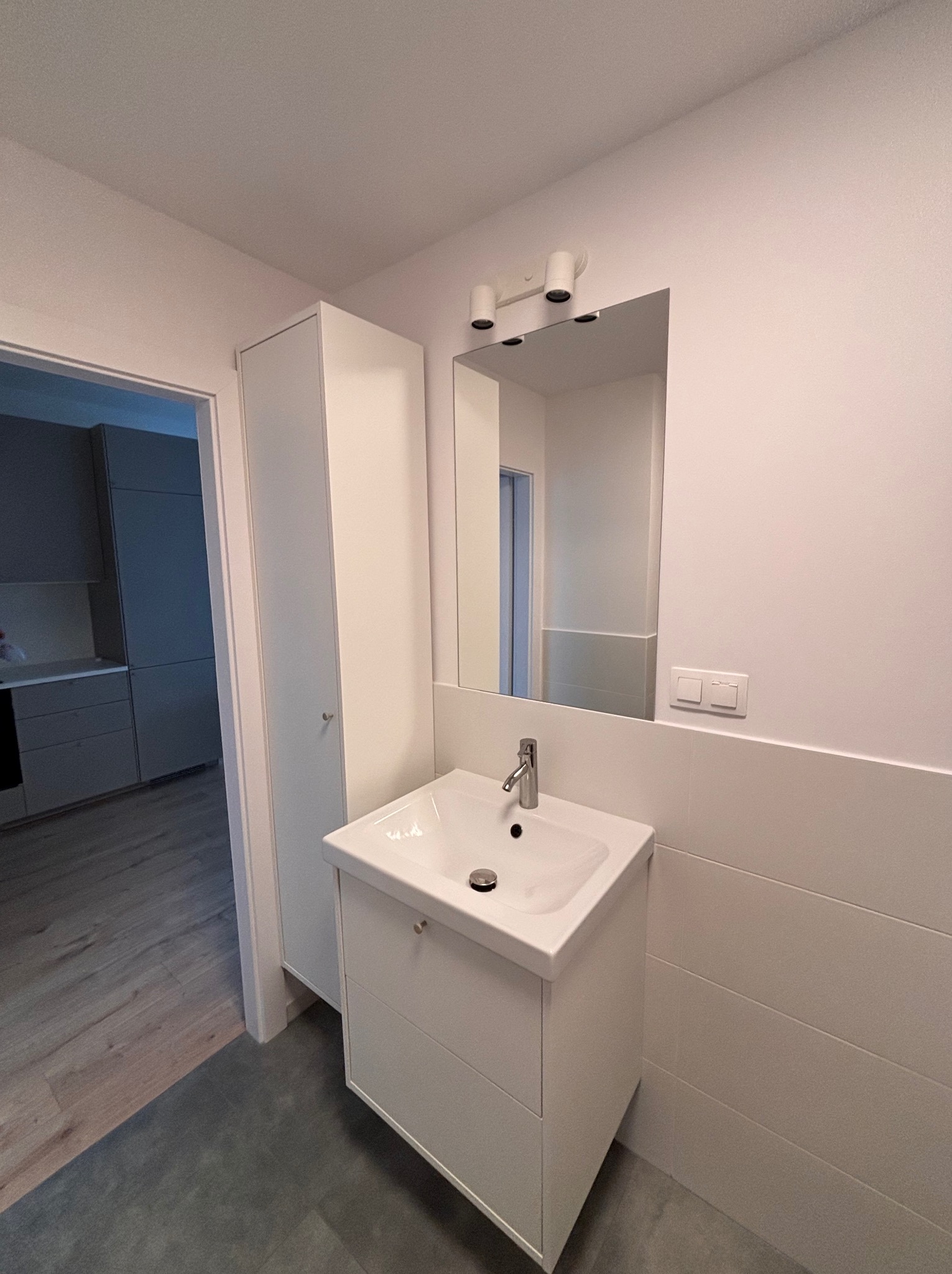 Biała umywalka z szafką i lustrem w nowoczesnej łazience. Widoczne płytki ścienne, oświetlenie nad lustrem i fragment kuchni w tle. Minimalistyczny design.