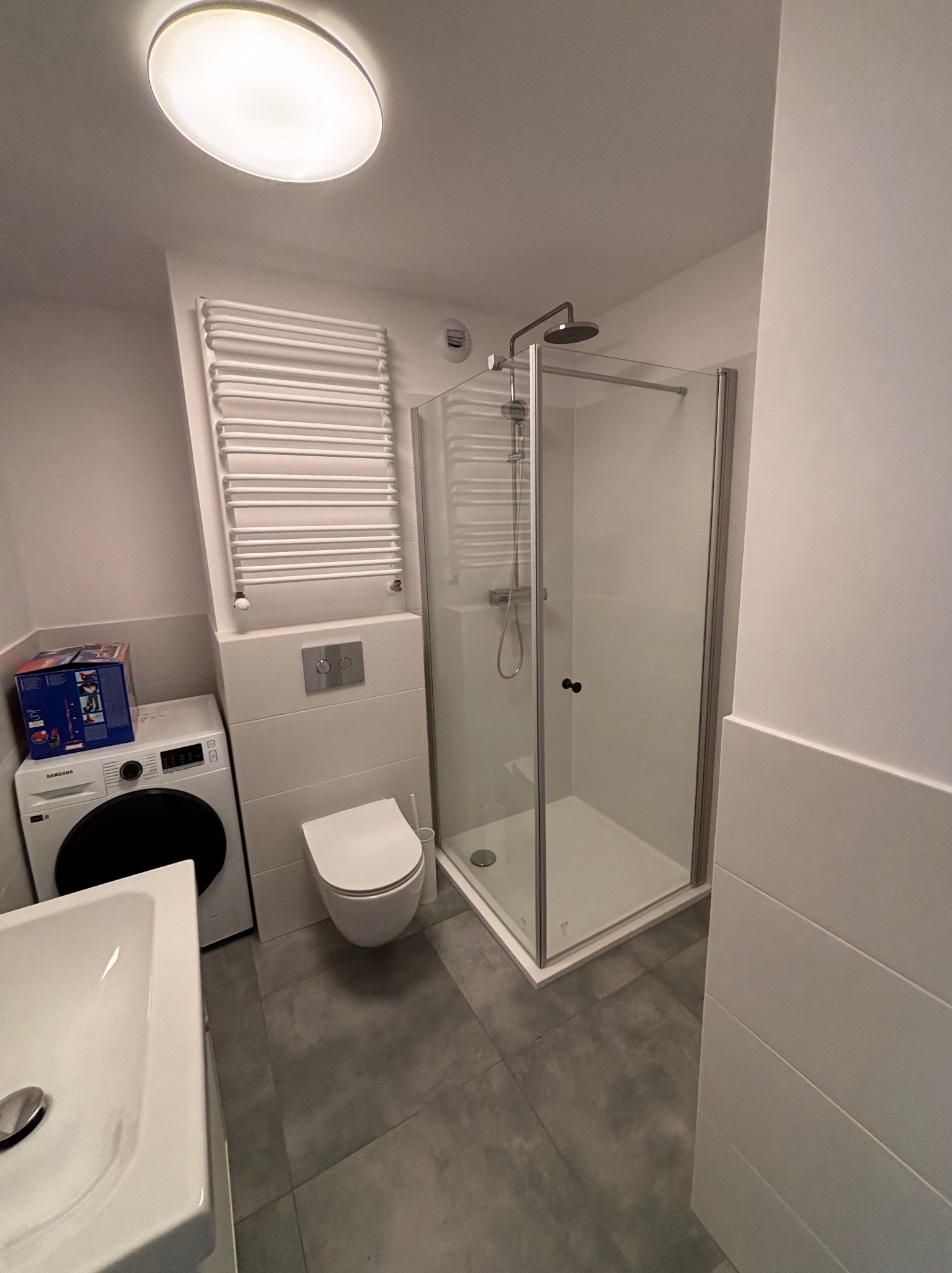 Nowoczesna łazienka po remoncie: kabina prysznicowa ze szklanymi drzwiami, podwieszana toaleta, grzejnik drabinkowy i pralka. Minimalistyczny design, szare płytki podłogowe.