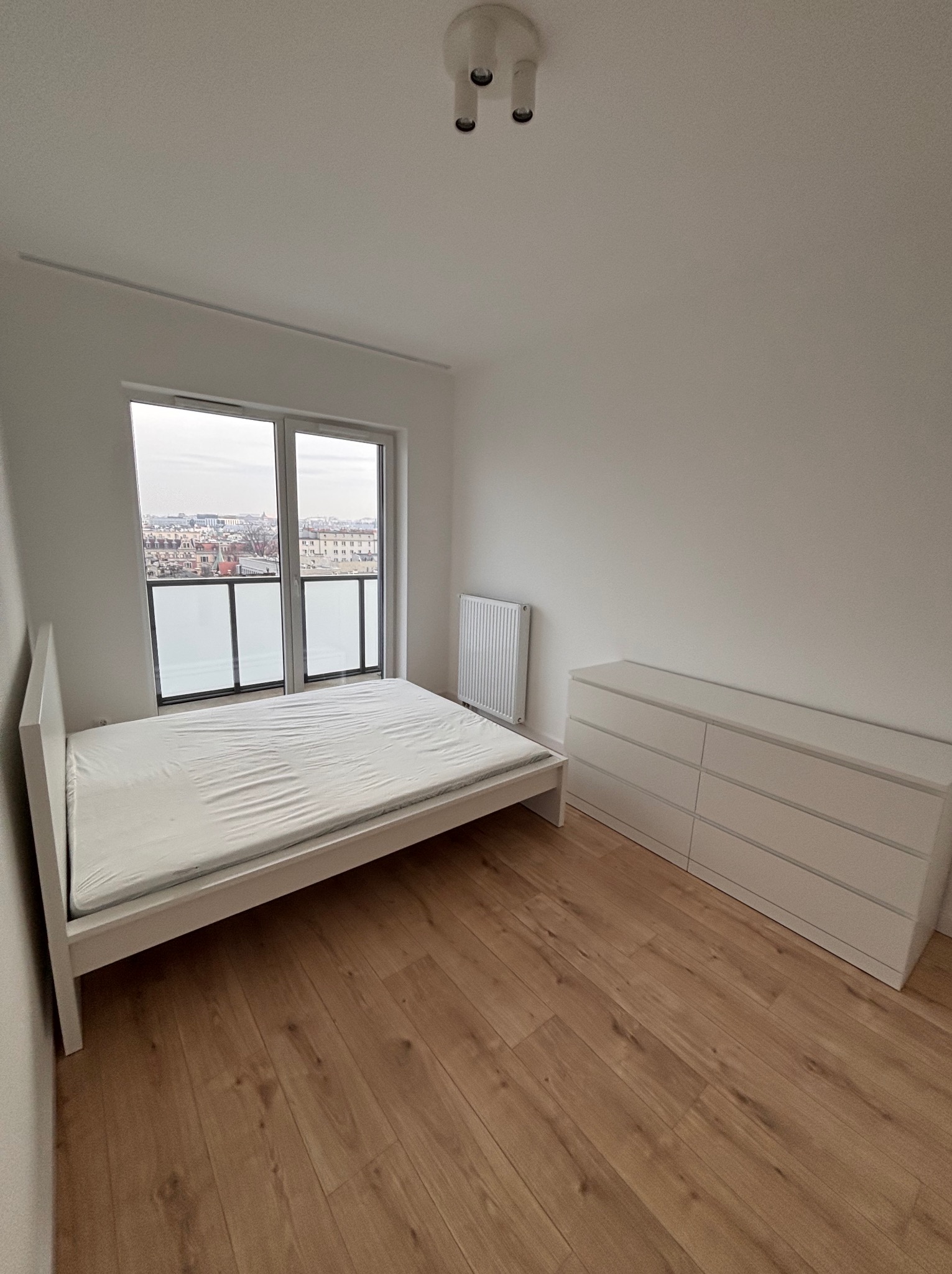Jasne wnętrze pokoju z drewnianą podłogą, białymi ścianami, łóżkiem, komodą i grzejnikiem. Widok na miasto przez okno. Minimalistyczny design.