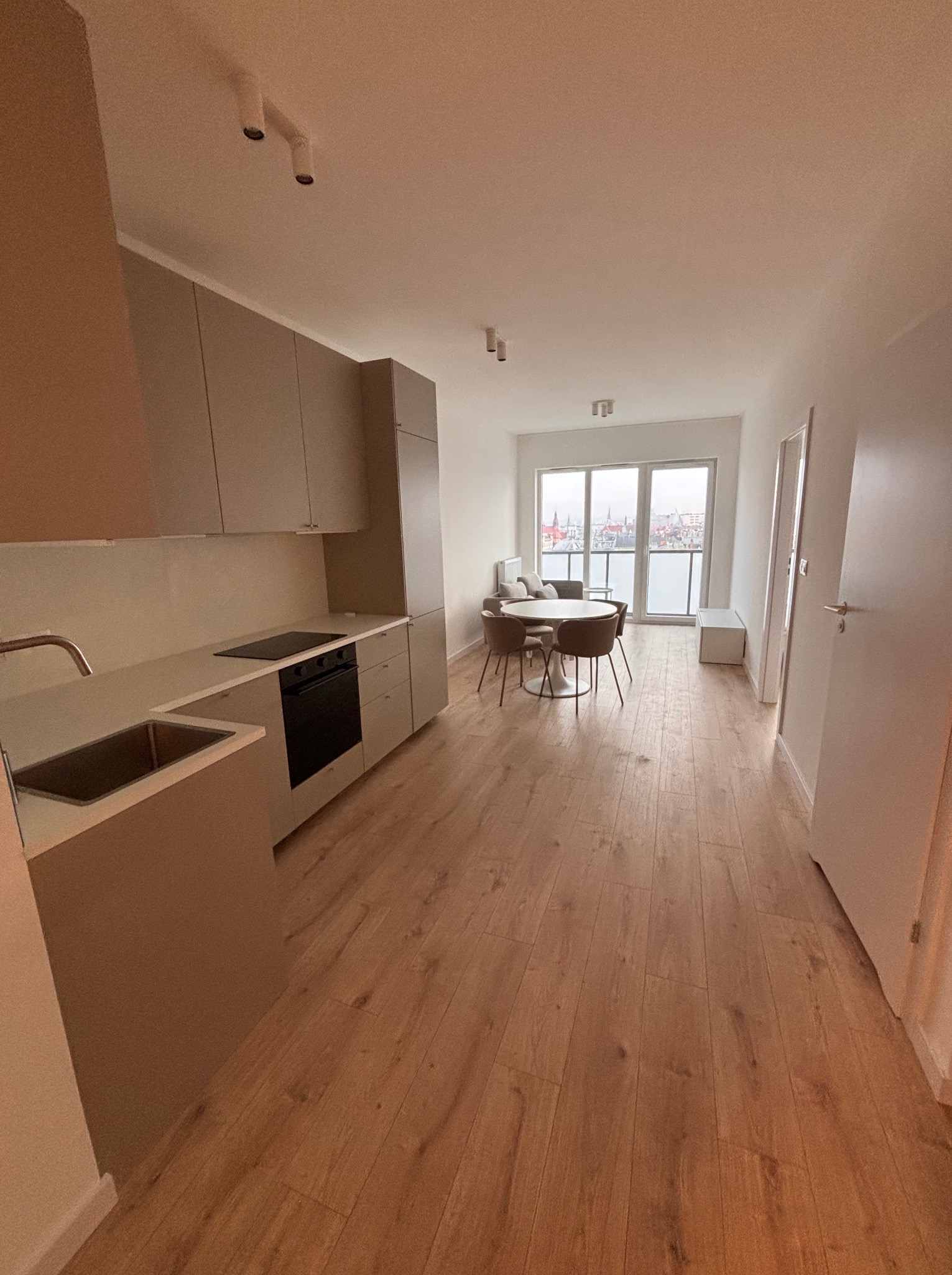 Jasne, przestronne wnętrze mieszkania po remoncie: drewniana podłoga, beżowa zabudowa kuchenna, okrągły stół z krzesłami, widok na miasto z balkonu. Minimalistyczny design.