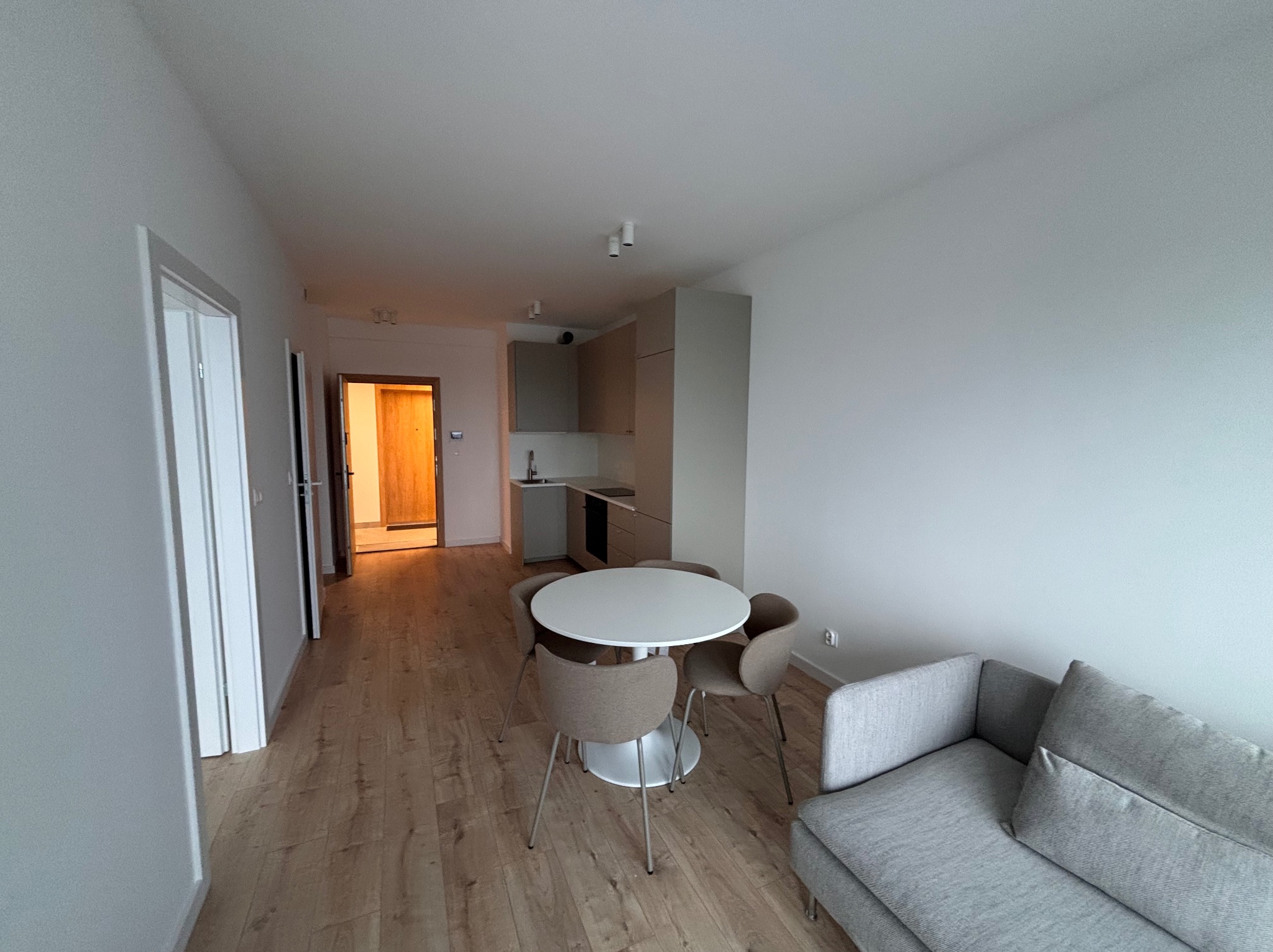 Jasne wnętrze mieszkania po remoncie: panele podłogowe, białe ściany, minimalistyczna kuchnia, okrągły stół z krzesłami i szara sofa. Widok na otwarte drzwi.