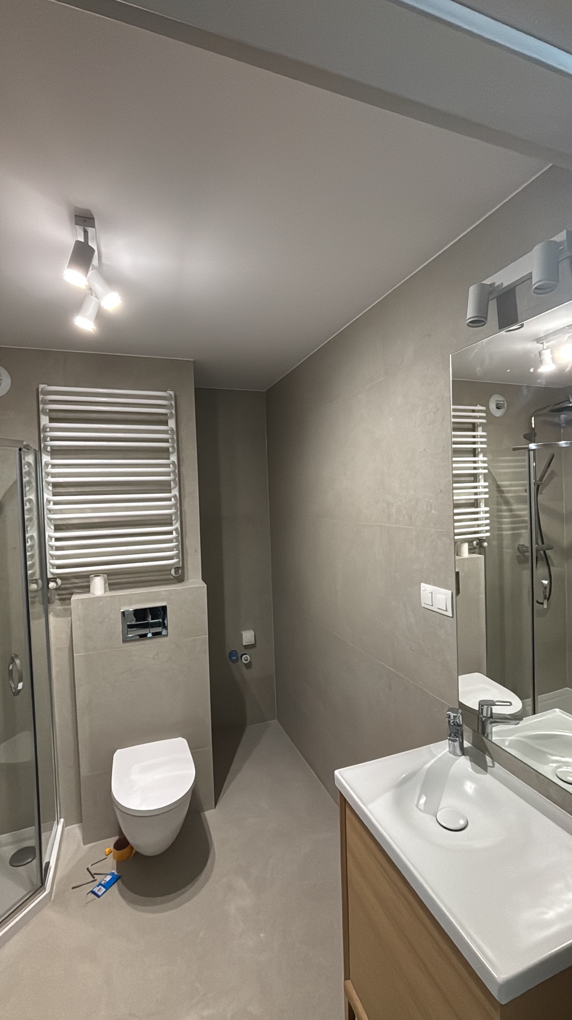 Nowoczesna łazienka w szarościach z białym wyposażeniem: toaleta, umywalka z szafką, grzejnik drabinkowy i kabina prysznicowa. Minimalistyczny design, oświetlenie punktowe.